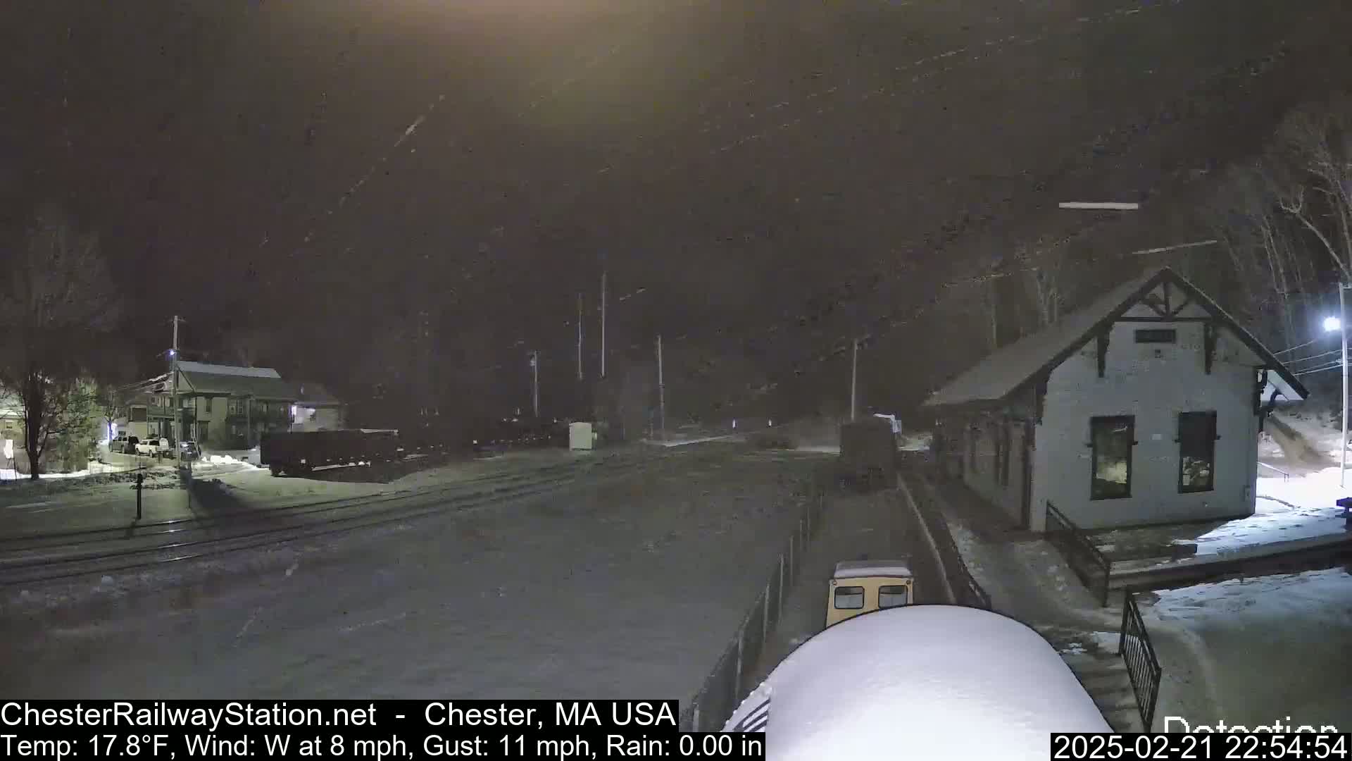 A snow-covered train approaches a small, snow-dusted train station at night in windy, cold weather.