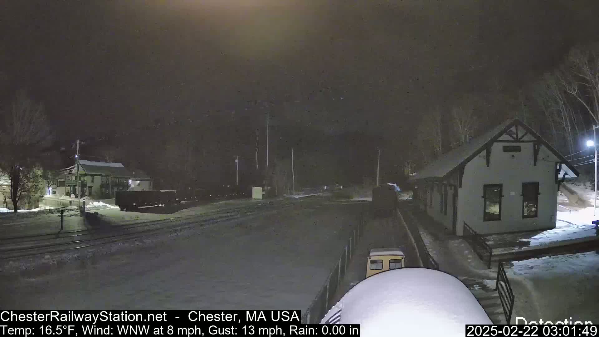 A snow-covered train yard at night, with a small building and rail cars visible under a dark sky.