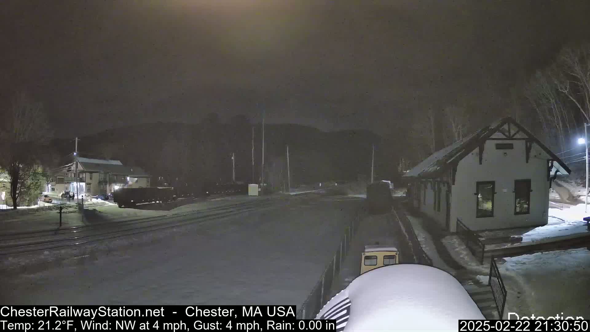A nighttime scene of a train station and rail yard covered in a light layer of snow, with a building to the right and train cars to the left, under a dark sky.