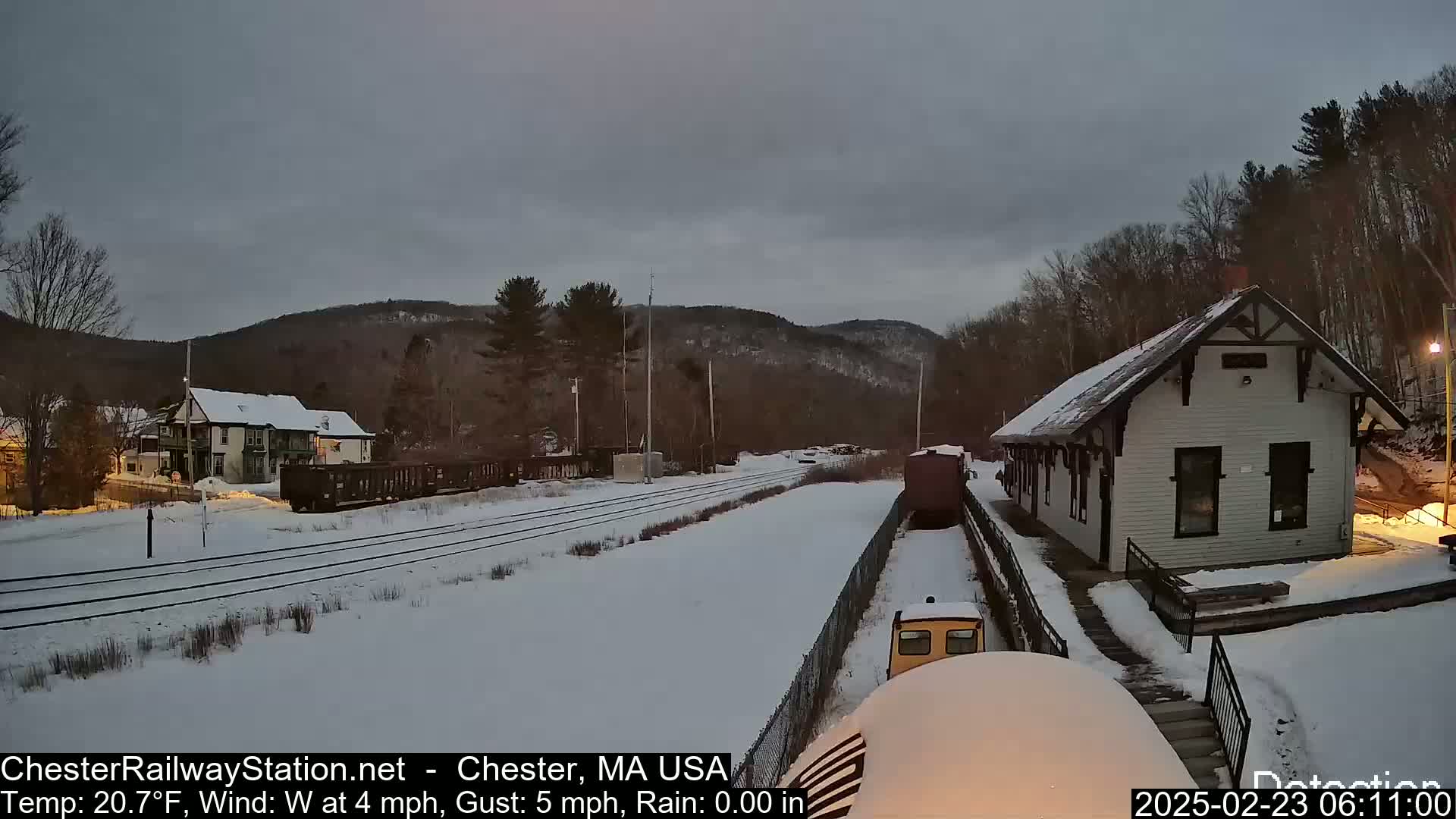 A snow-covered train station and railway tracks are visible under a gray sky, with a train partially visible near the station.