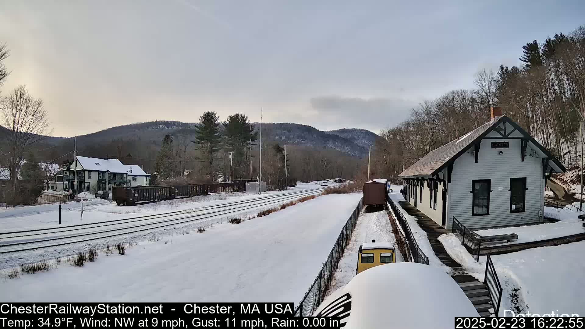 A snow-covered train station and rail yard are situated in a valley, with a partly cloudy sky and a temperature of 34.9°F.