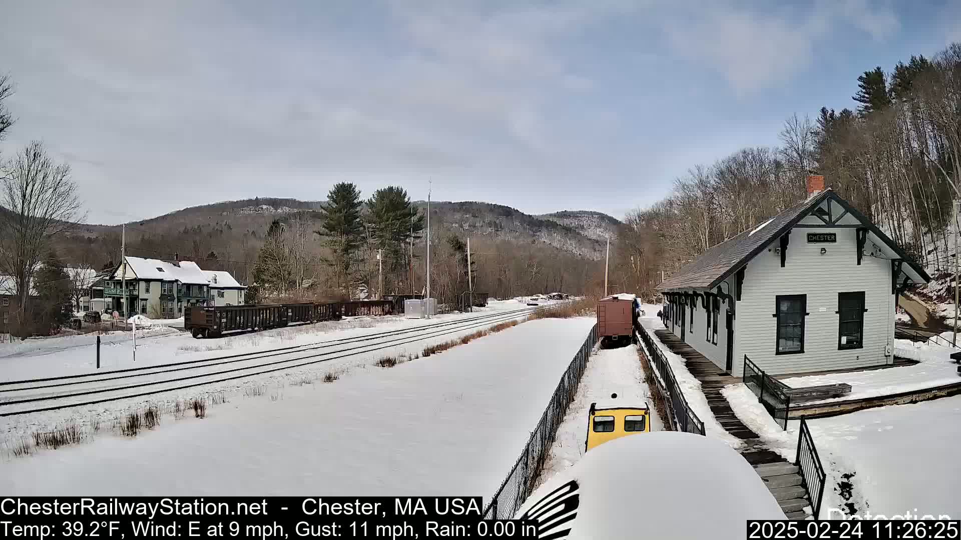 A snow-covered train station and tracks sit nestled in a wooded valley under a partly cloudy sky.