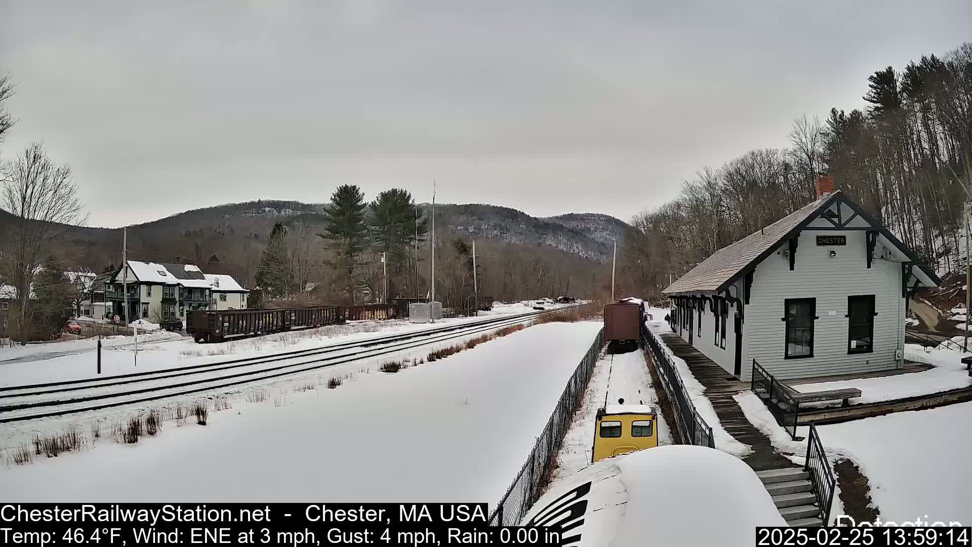 A snow-covered train station and rail yard with a train approaching, under an overcast sky.