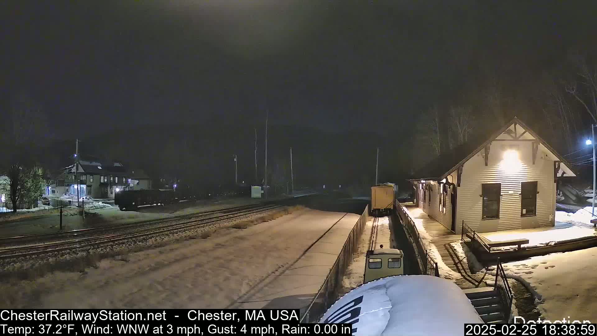 A nighttime view of a snow-covered train station and railway tracks with a few train cars visible, under a dark sky with a wind from the west-northwest.
