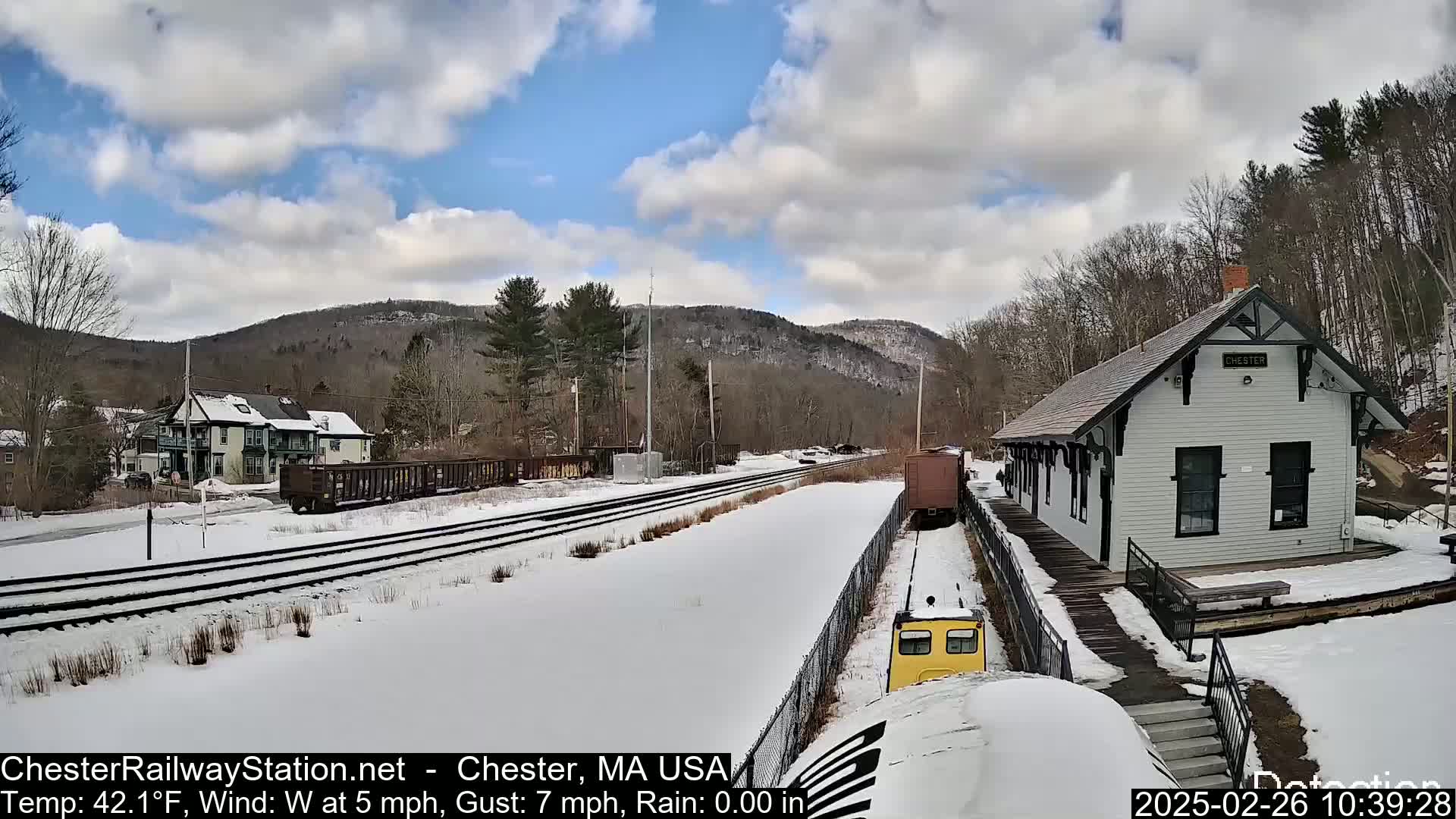 A snow-covered train station and tracks are visible, with a small yellow train car approaching, under a partly cloudy sky.