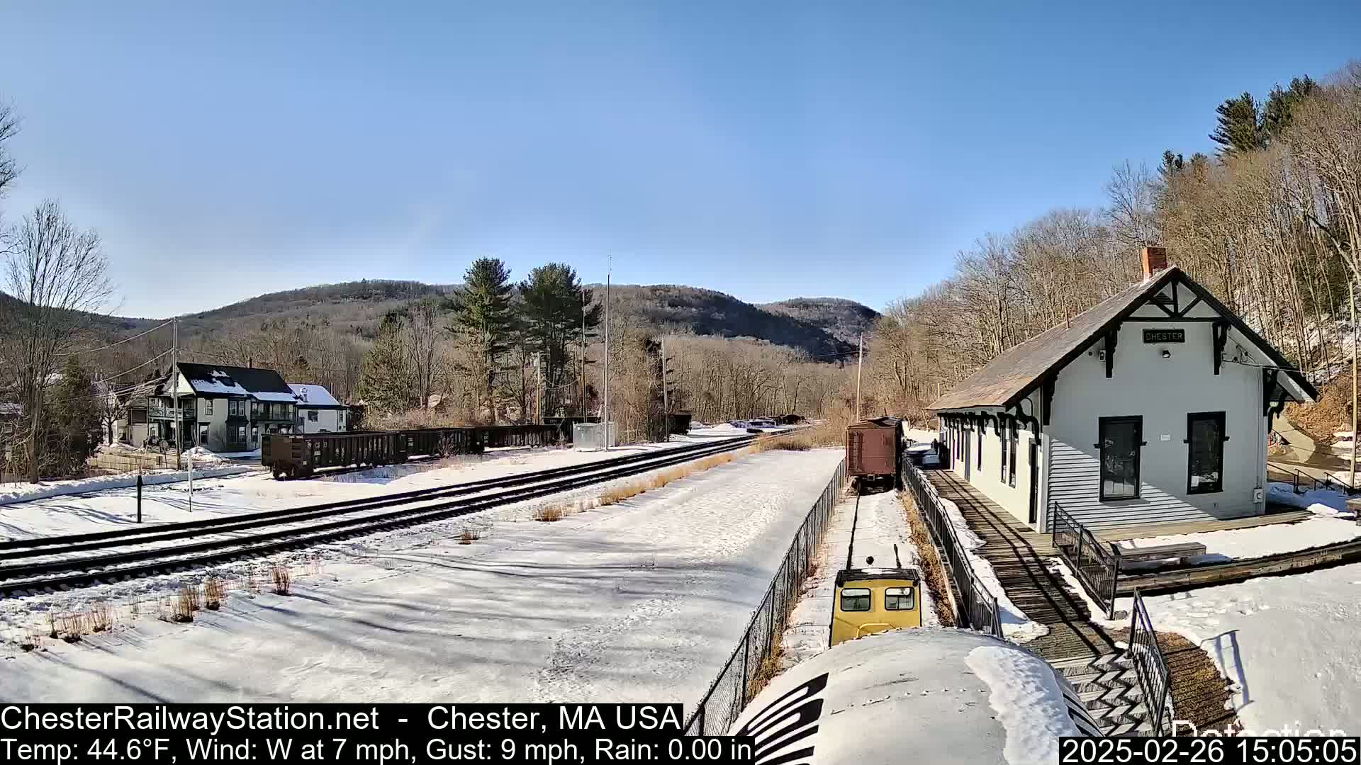 A snow-covered train station and tracks are situated in a valley, with a partly sunny sky and bare trees.