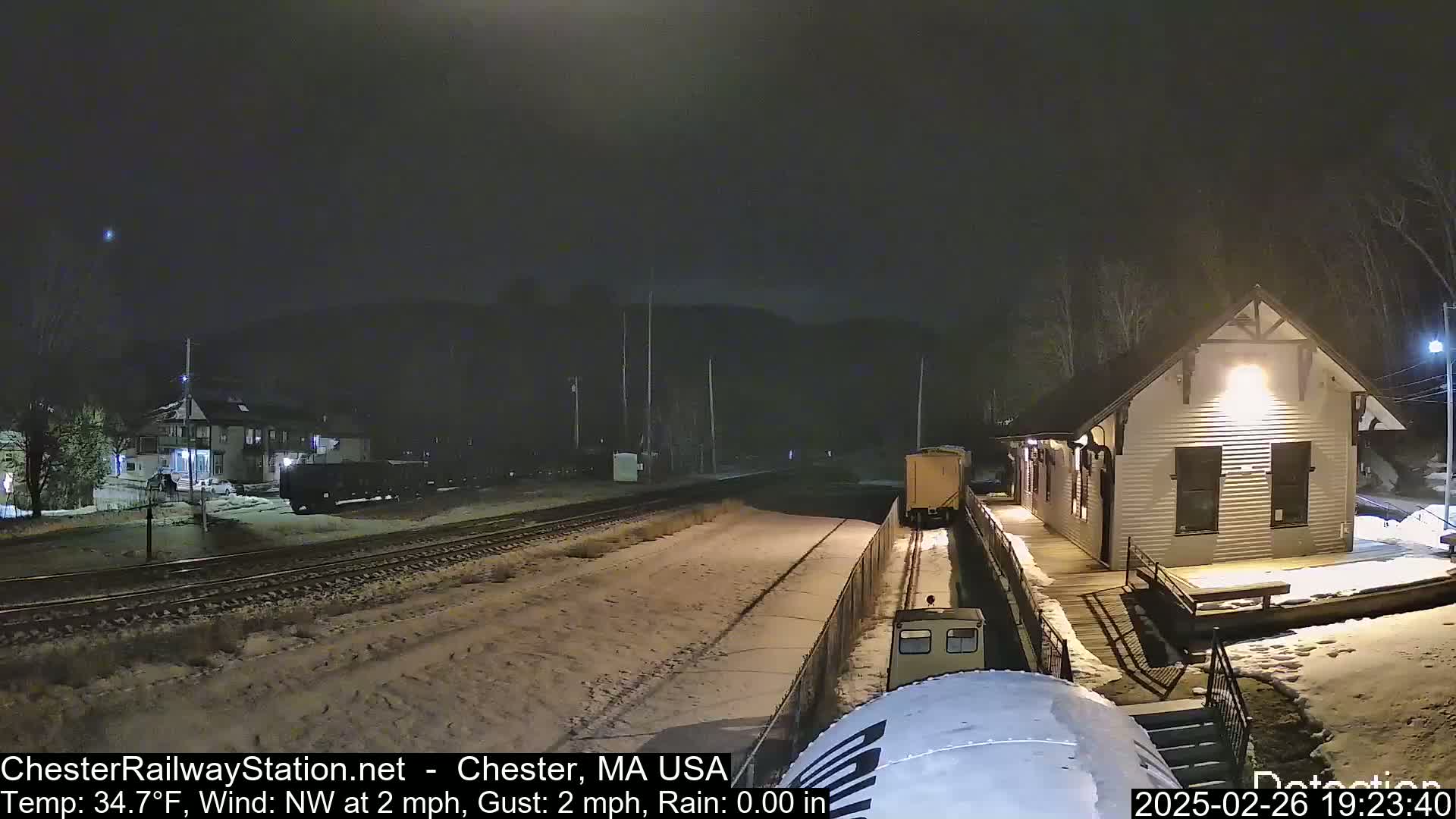 A snow-covered train station at night is illuminated, with a train partially visible on the tracks in front of it, under a dark sky with a northwest wind.