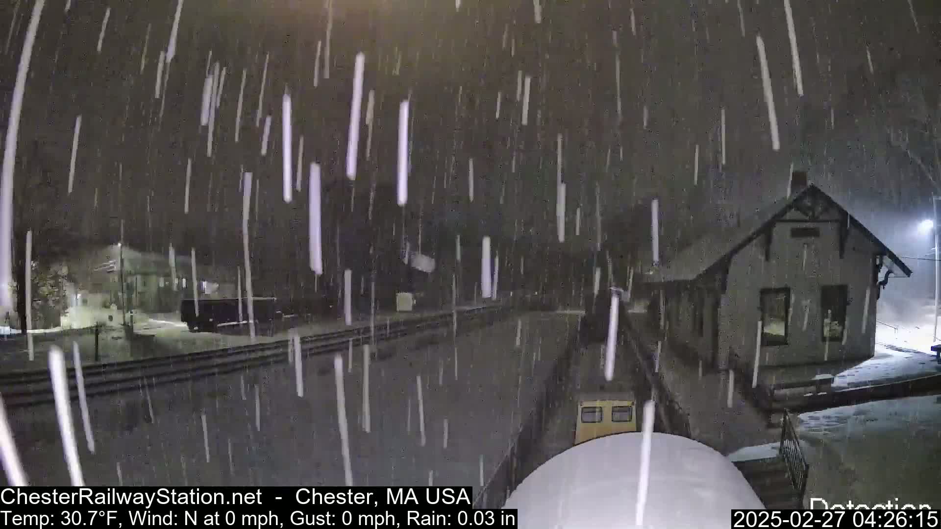 A nighttime scene of a train station covered in snow during a light rain, with a train partially visible in the foreground.