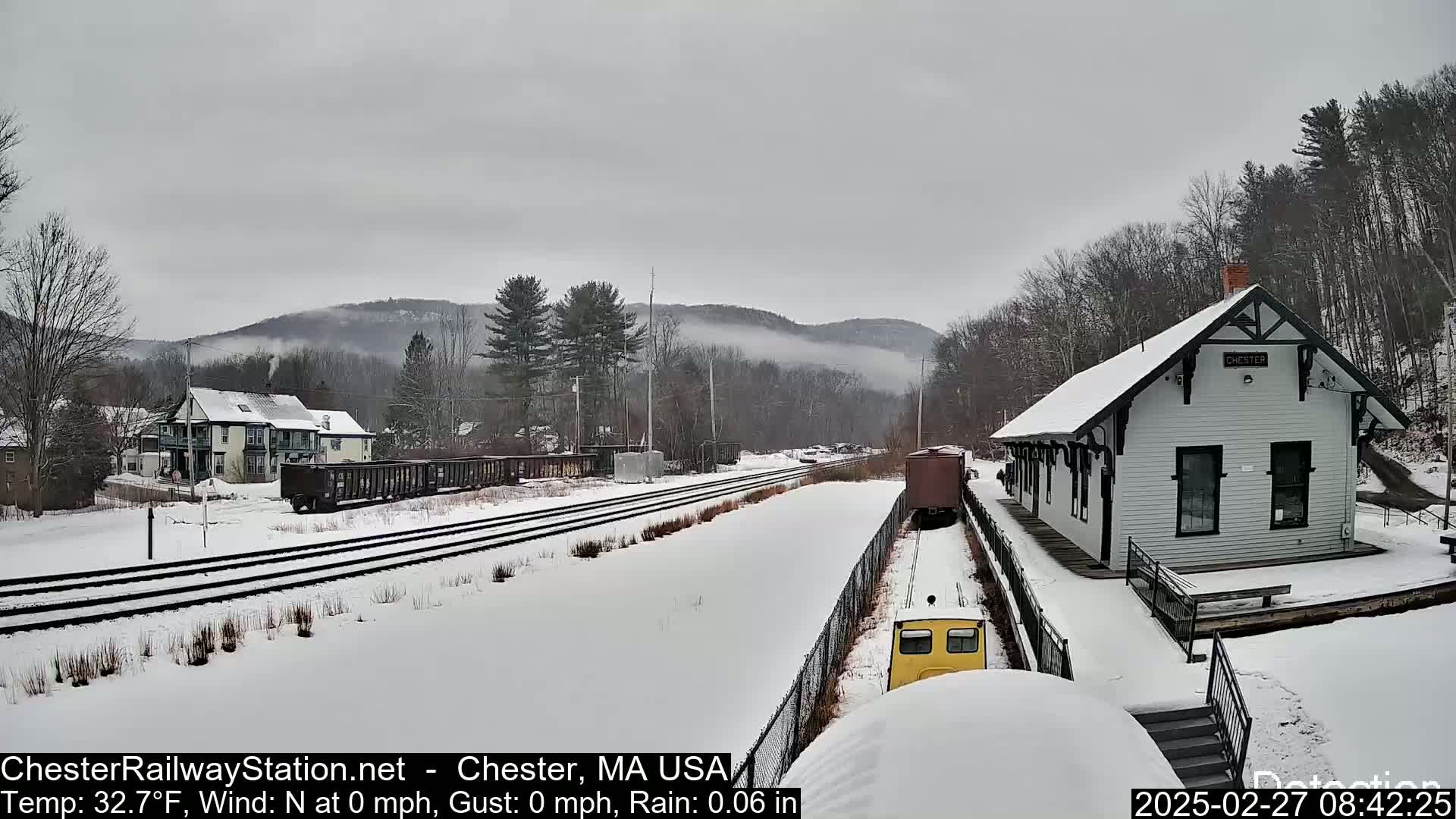 A snow-covered train station sits beside a railway track in a lightly snowing landscape, with misty hills in the background.