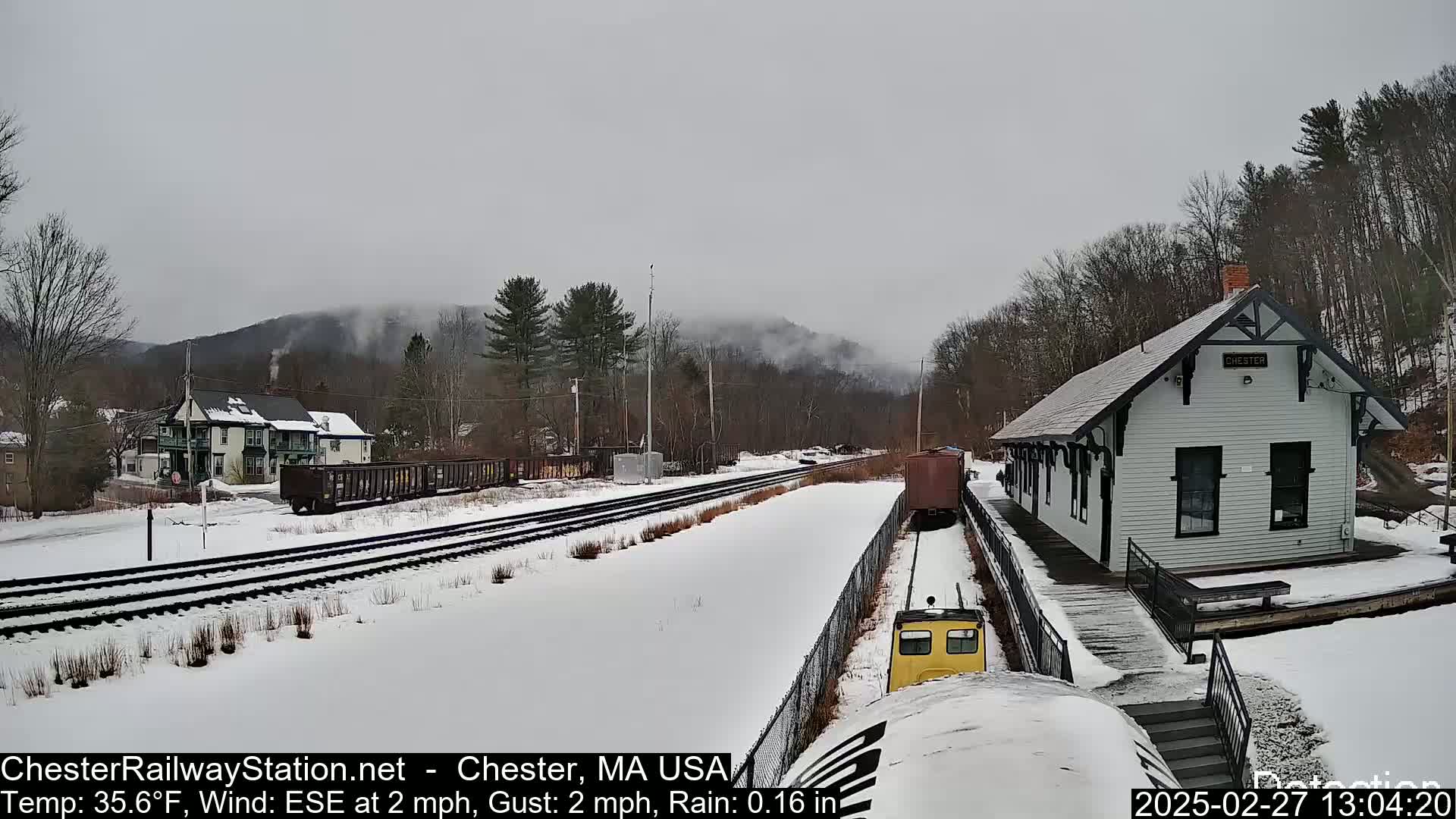 A snow-covered railroad scene shows a train approaching a small, white station building under a gray, rainy sky.