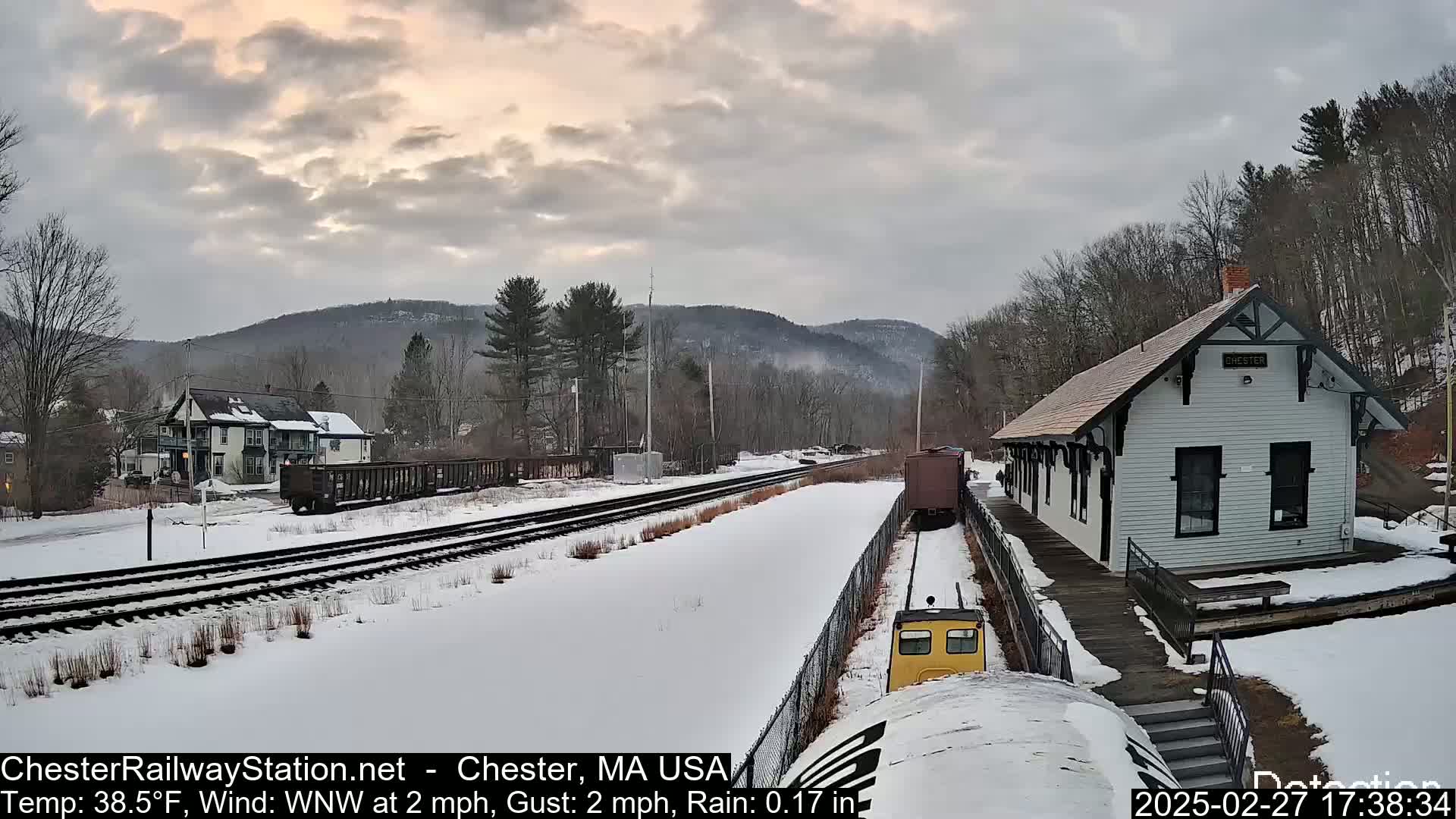 A snow-covered train station and tracks are visible, with a train car approaching the station under a cloudy, twilight sky.