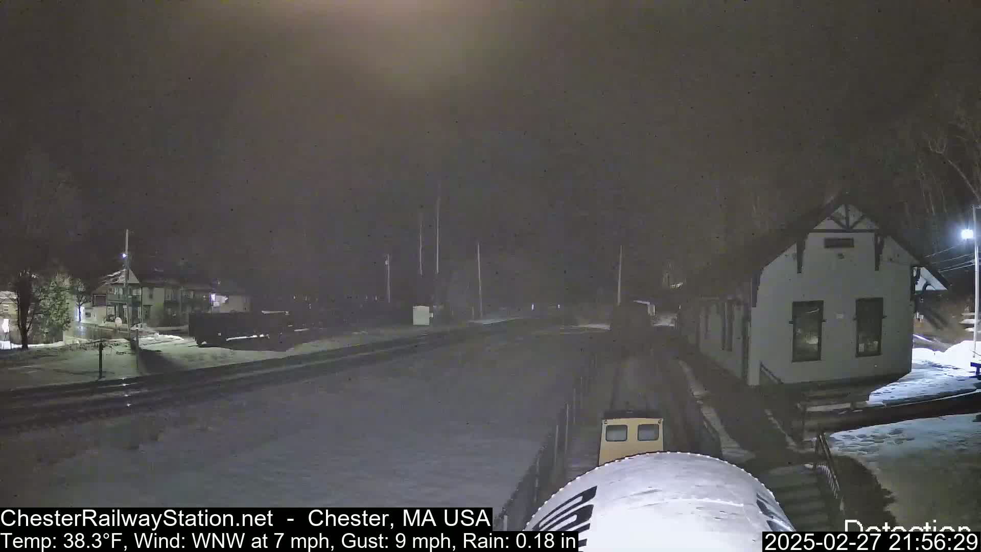 A nighttime view of a snow-covered train station and surrounding area under rainy conditions with a wind from the west-northwest.