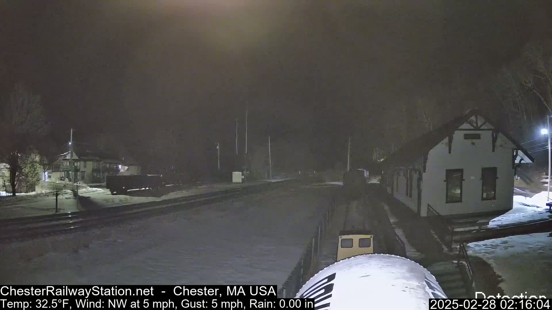 A nighttime view of a snow-covered train station and railroad tracks under a dark, cloudy sky with a northwest wind.