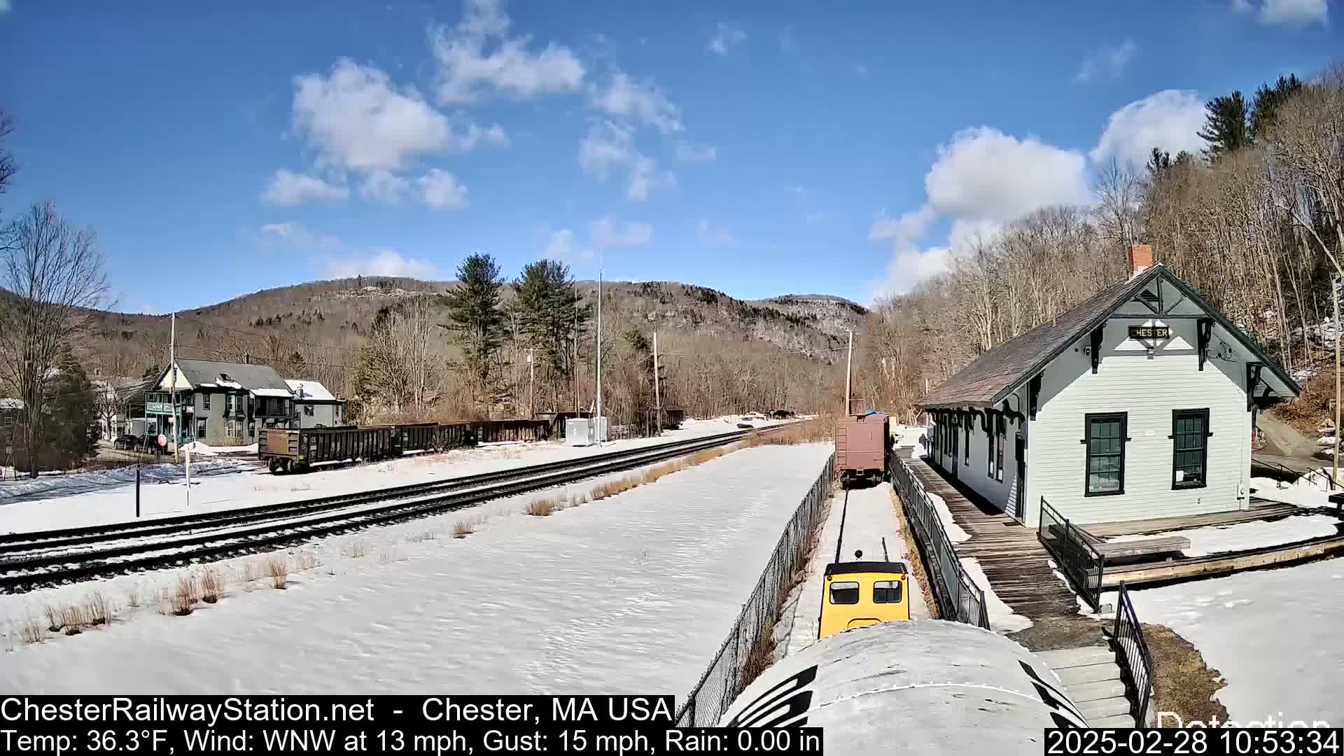A snow-covered train station and rail yard with a small yellow train in the foreground, under a partly cloudy blue sky.