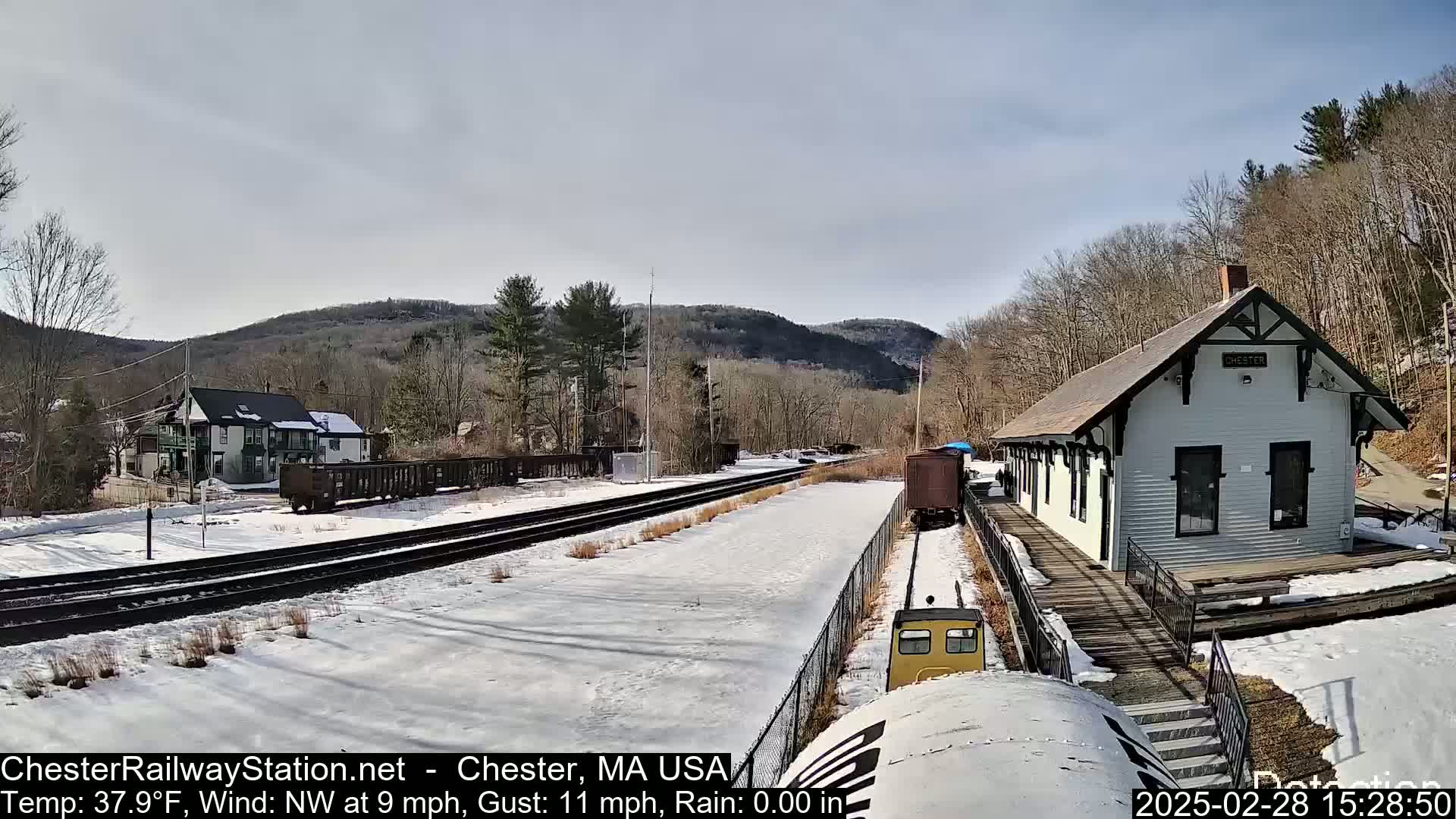 A snow-covered train station and tracks are nestled in a valley between leafless trees and hills under a partly sunny sky.