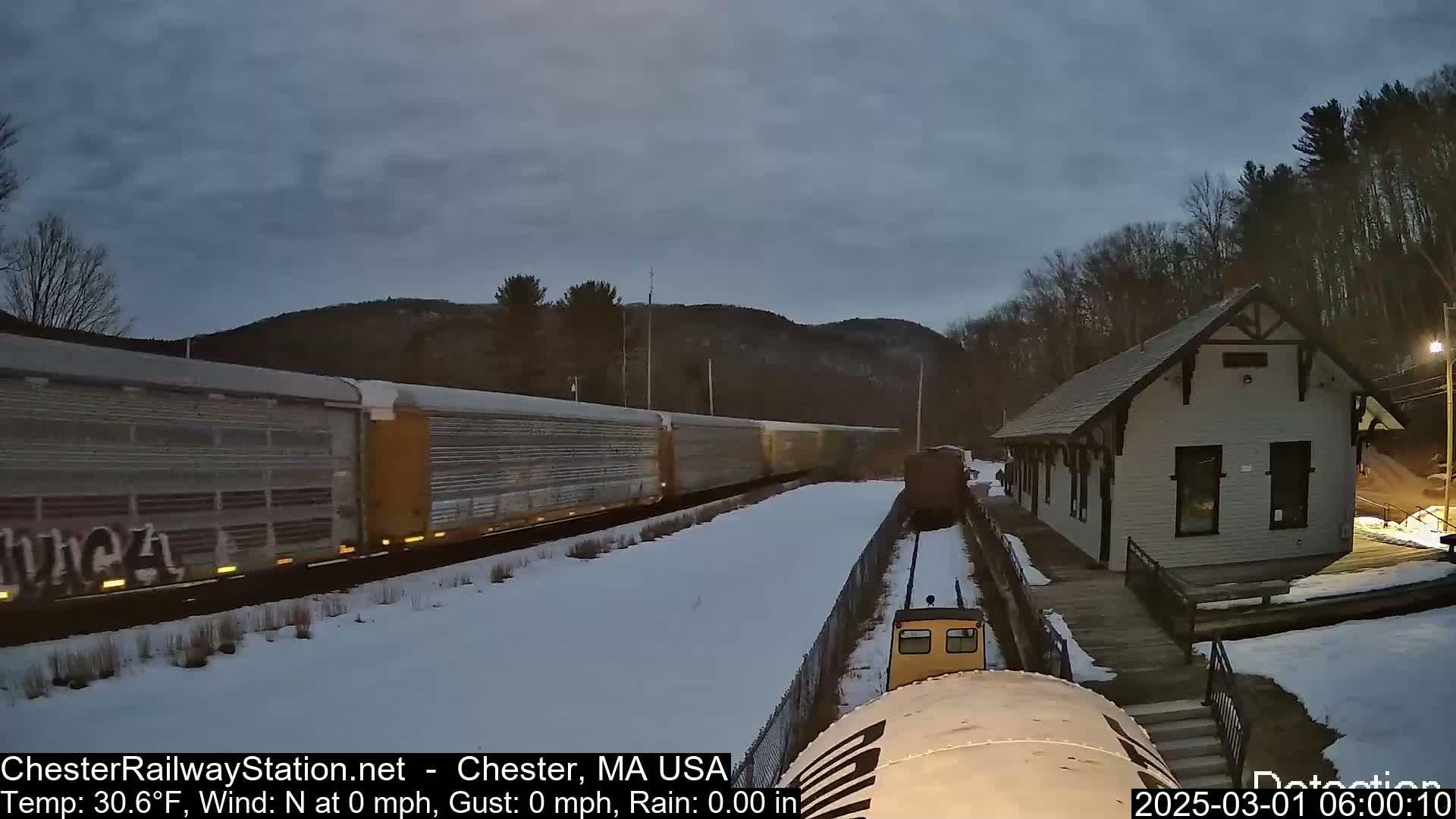 A long freight train passes a snow-covered train station under a twilight sky.