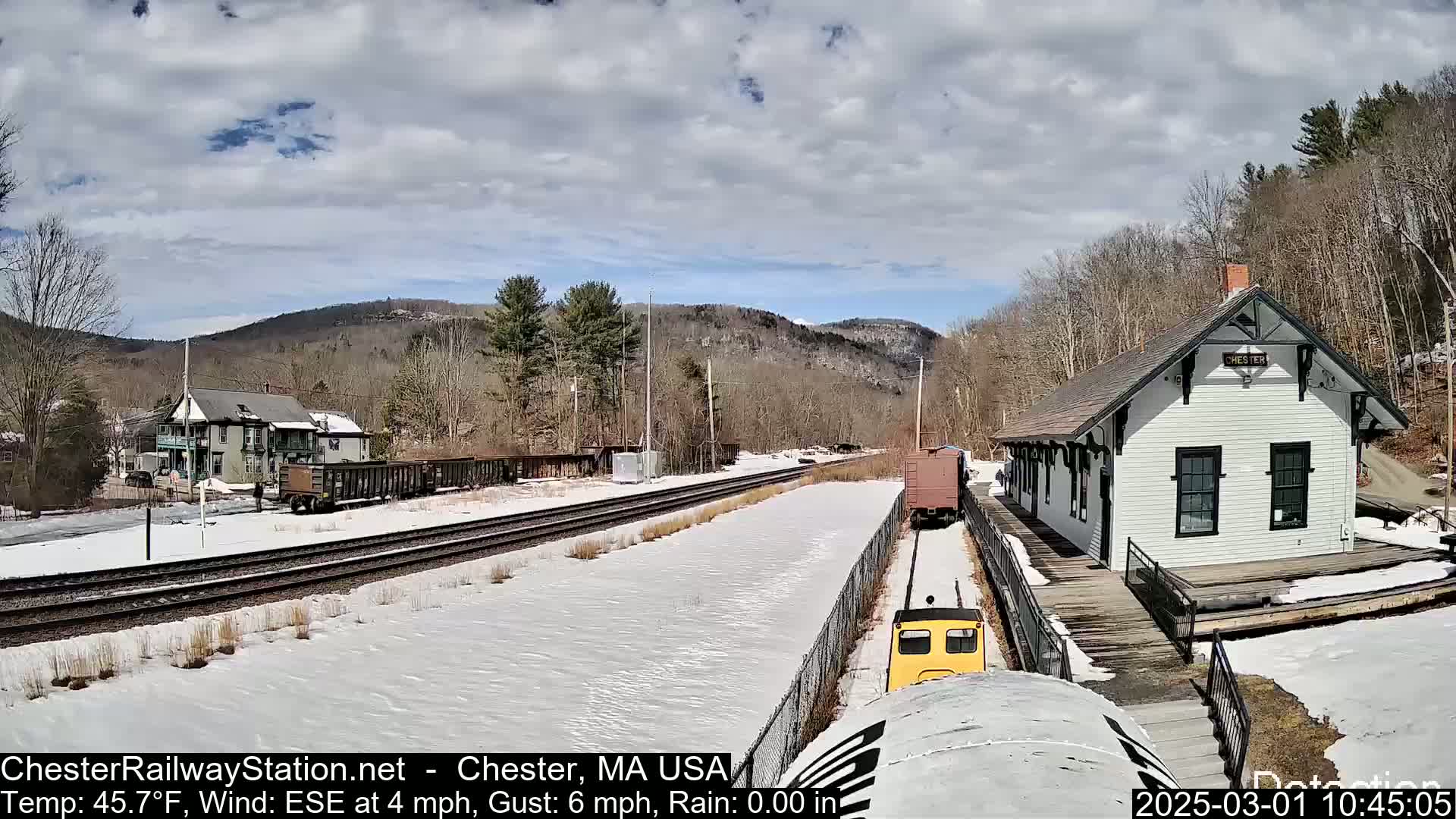 A yellow train approaches a snow-covered train station in a partly sunny, cold setting with mountains in the background.