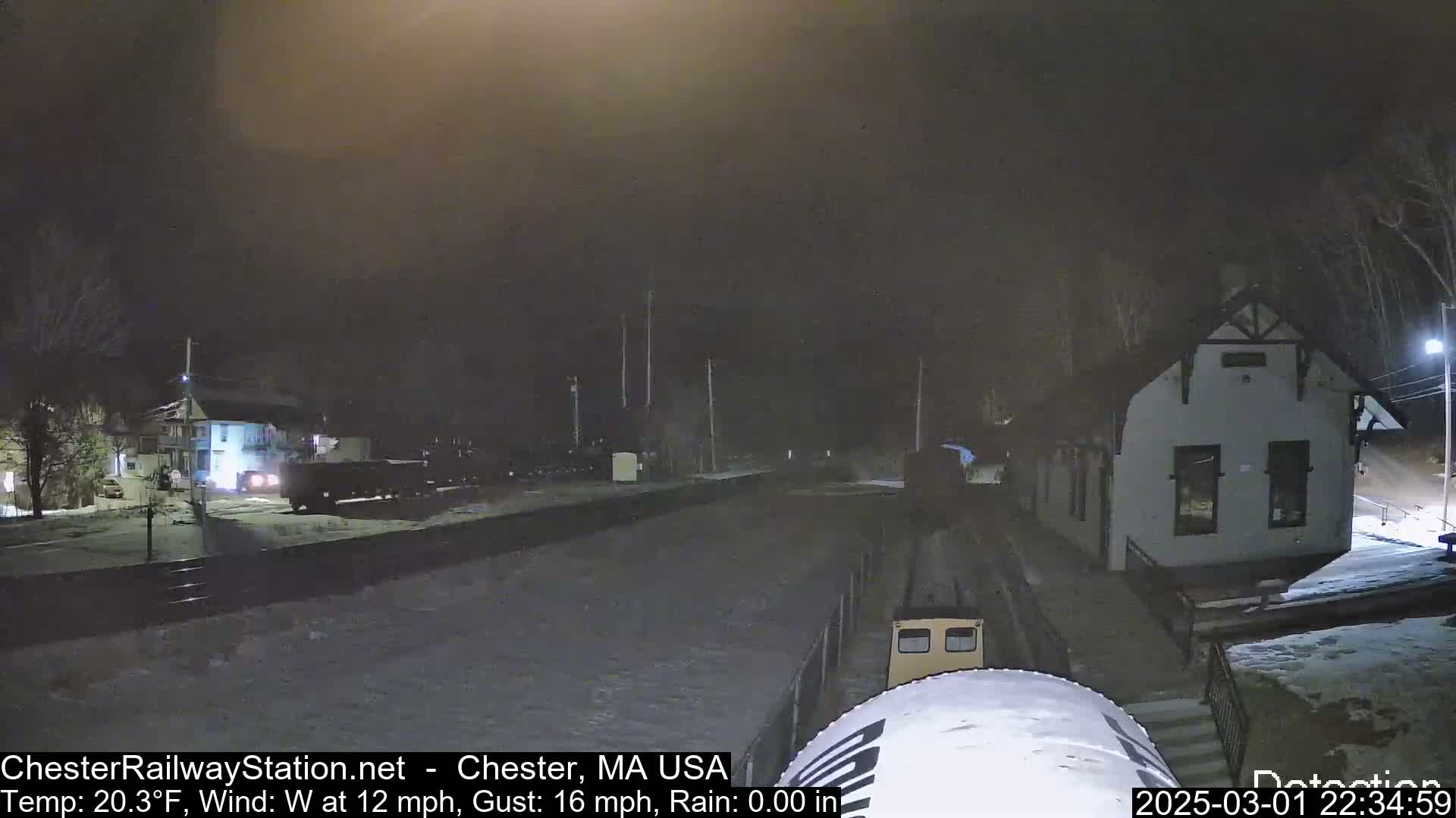 A nighttime scene of a train approaching a snow-covered station under a dark, hazy sky with a 20.3°F temperature, a 12 mph west wind, and 16 mph gusts.