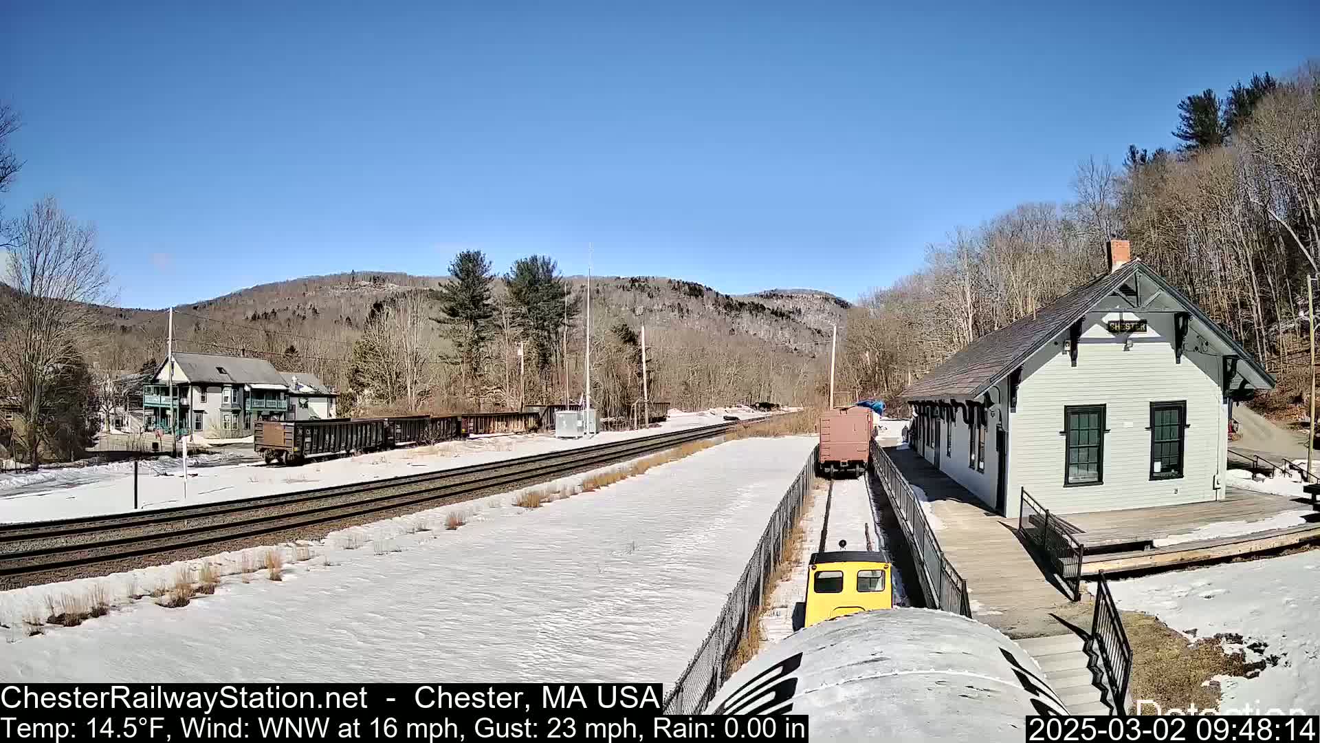 A small yellow train approaches a light-grey train station on a sunny day with snow on the ground, and train cars are visible in the background.