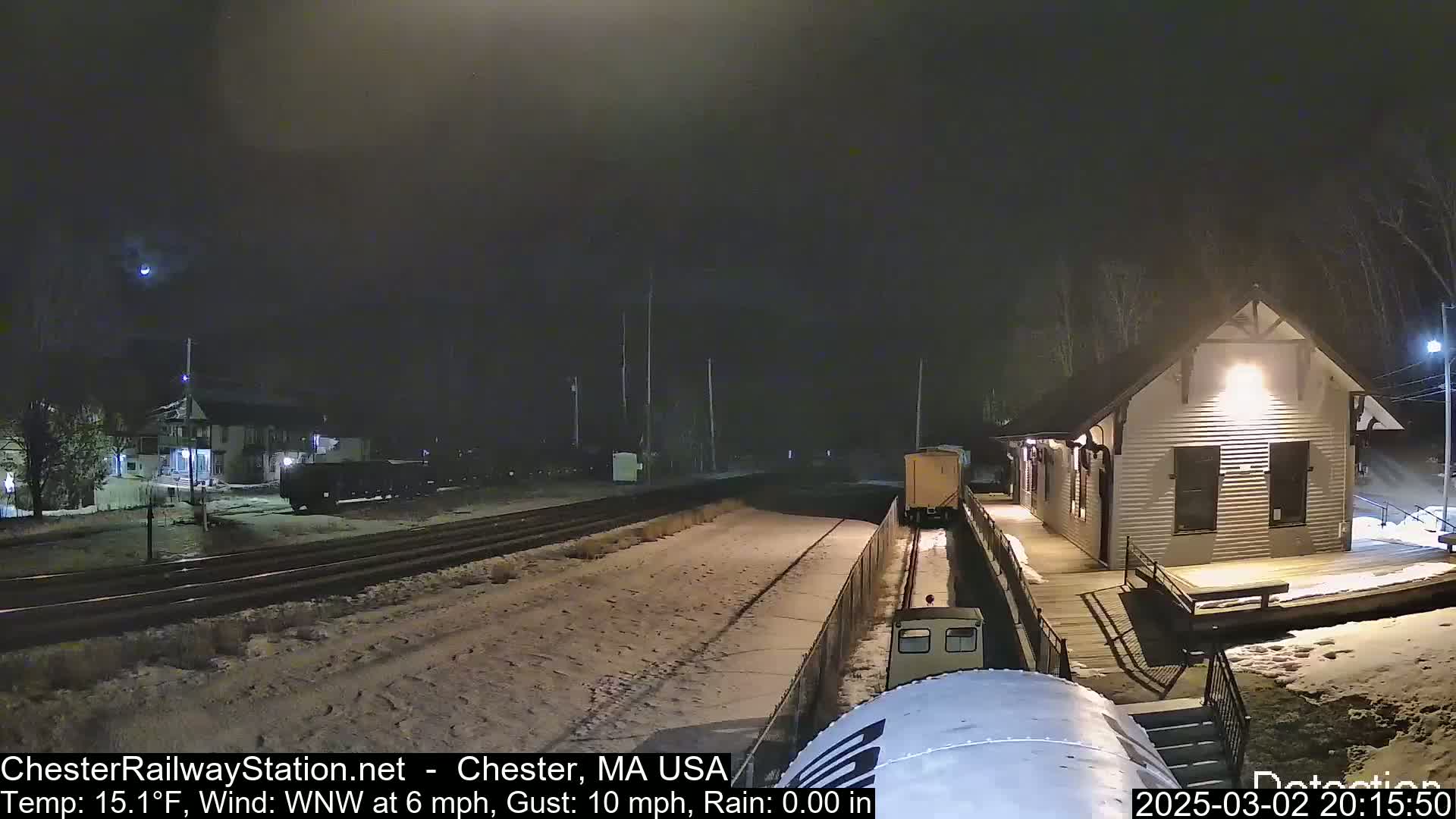 A train passes a snow-covered train station at night in windy, cold conditions.