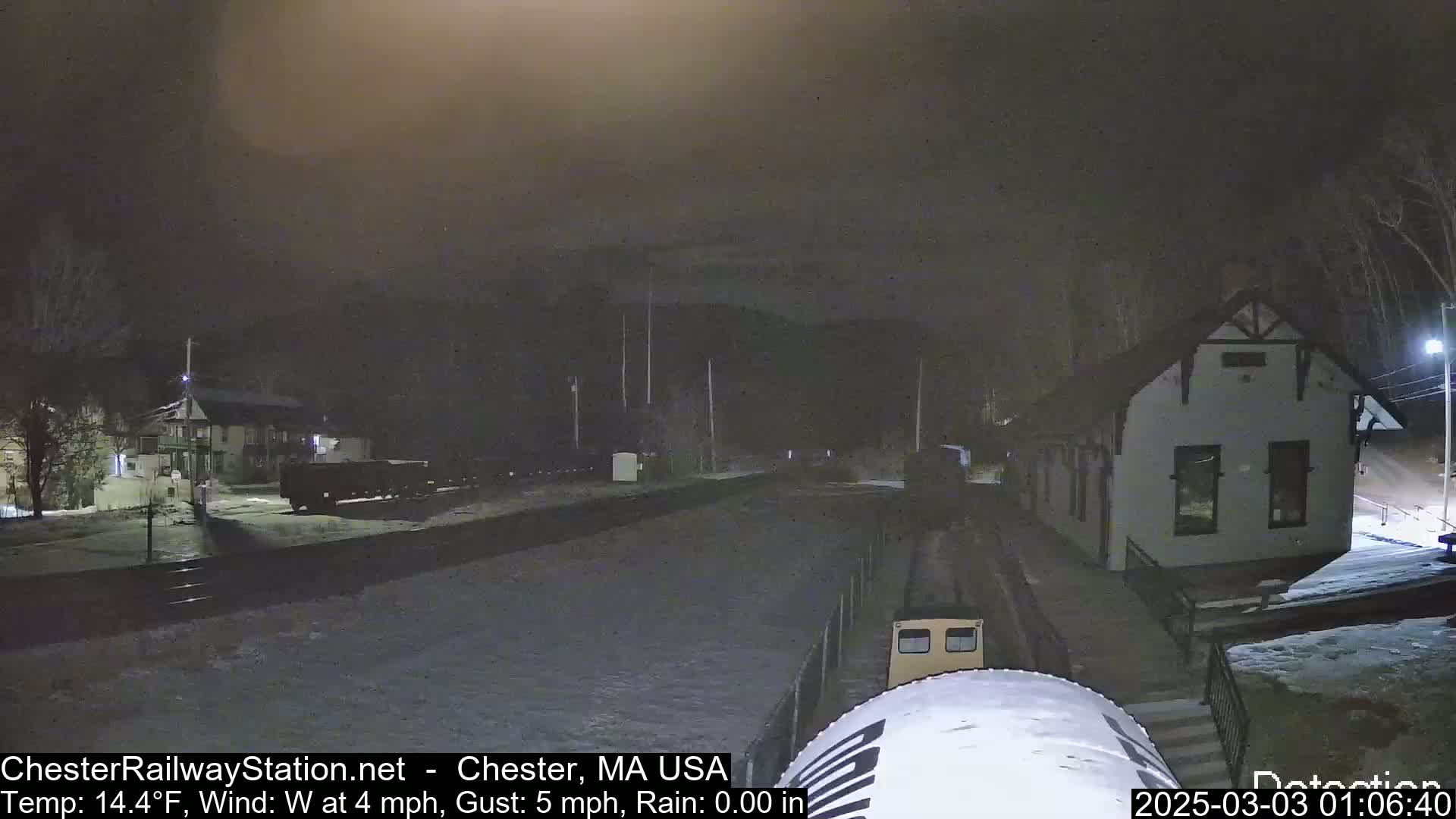 A nighttime view of a train approaching a station building under a dark, overcast sky, with snow or ice on the ground.