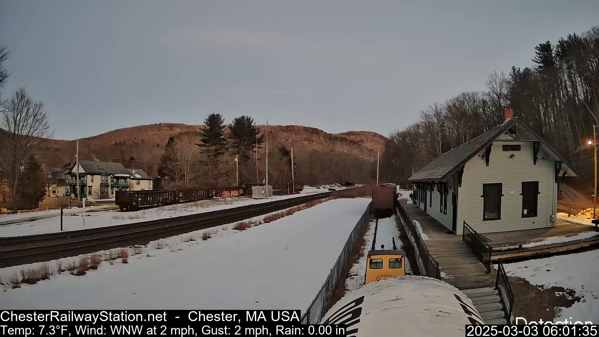 A snow-covered train station and tracks are visible under a twilight sky, with a small yellow train car approaching the station.