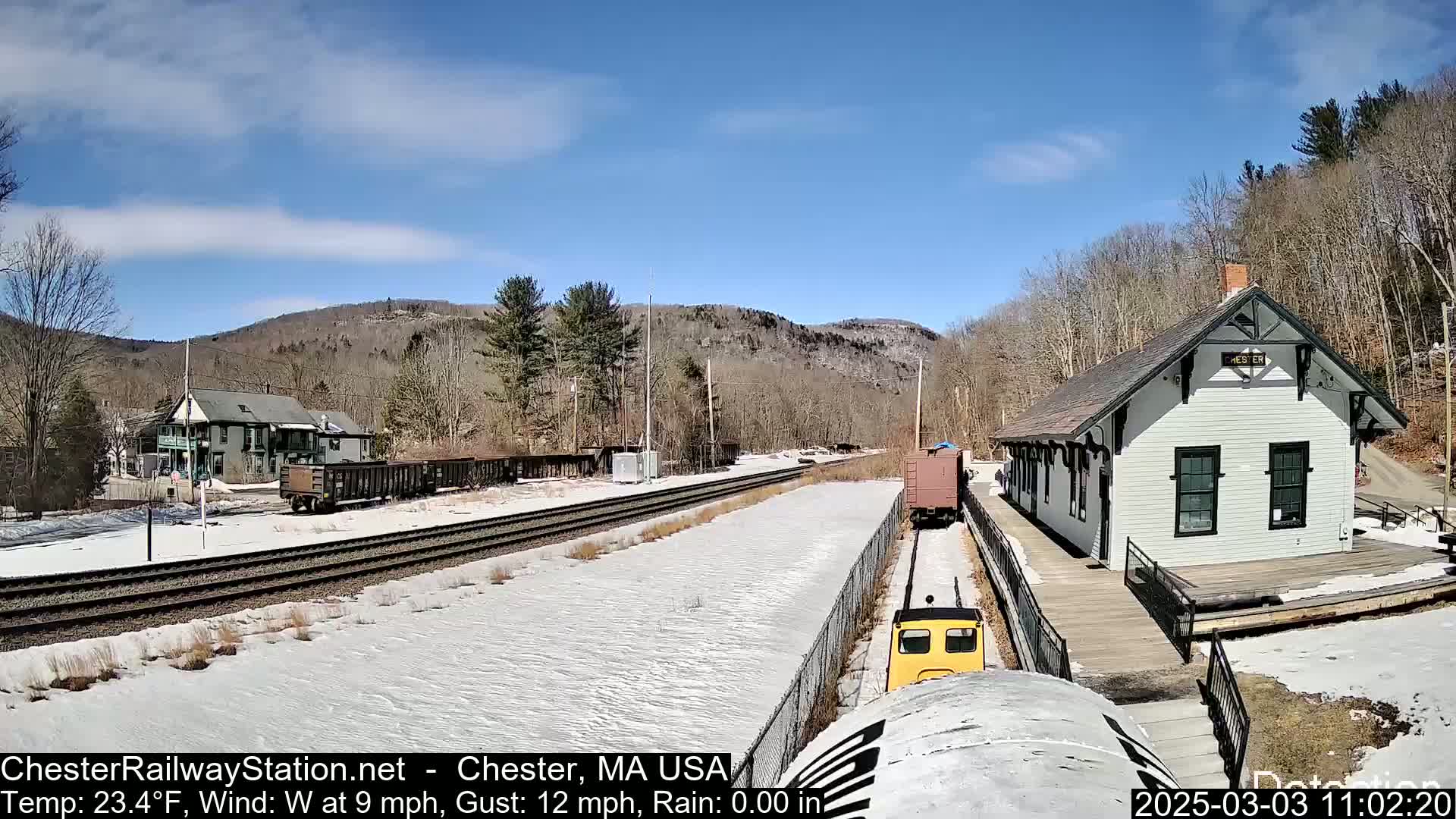 A small yellow train approaches a snow-covered train station on a sunny day with a light westerly wind.