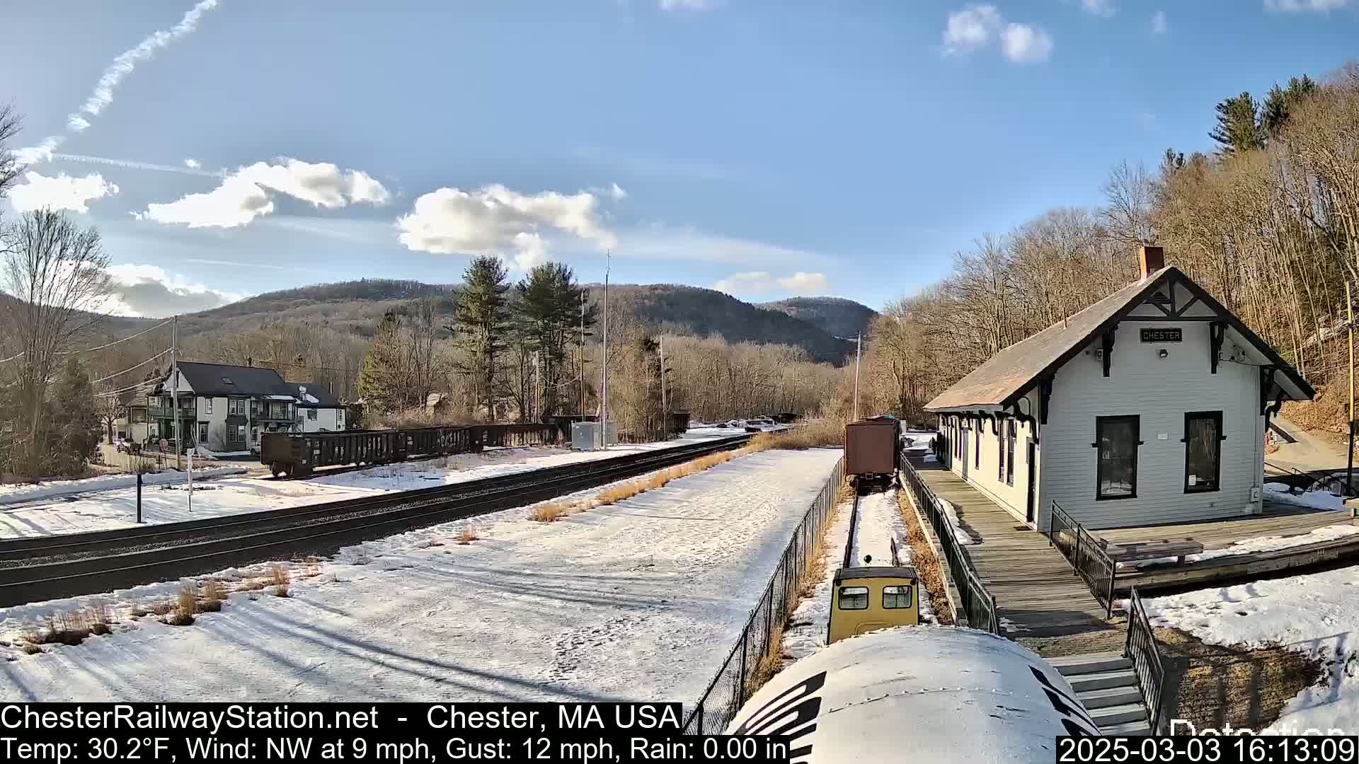 A snow-covered train station and rail yard are nestled among bare trees and hills under a partly sunny sky.