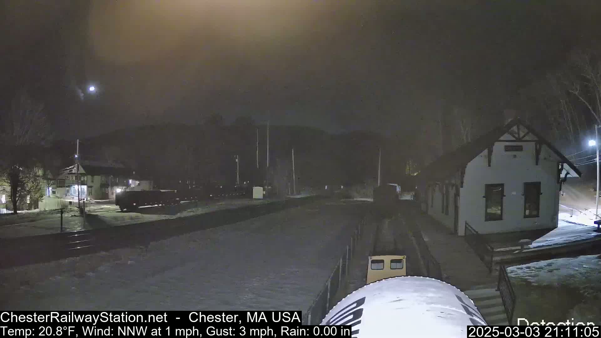 A nighttime view of a snow-covered train station and tracks under a dark, cloudy sky, with a light rain and a temperature of 20.8°F.