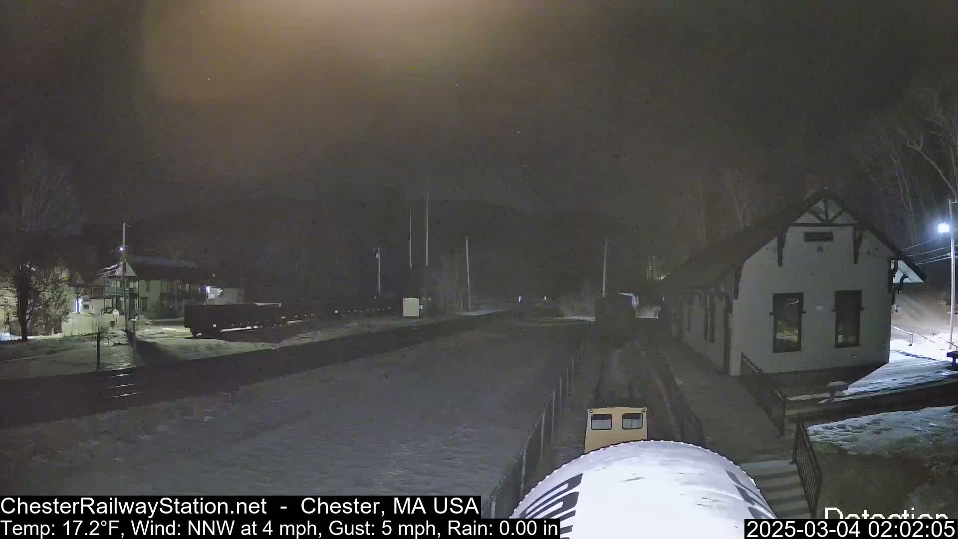 A nighttime scene of a train partially visible near a station building, in the snow, under a dark, overcast sky.