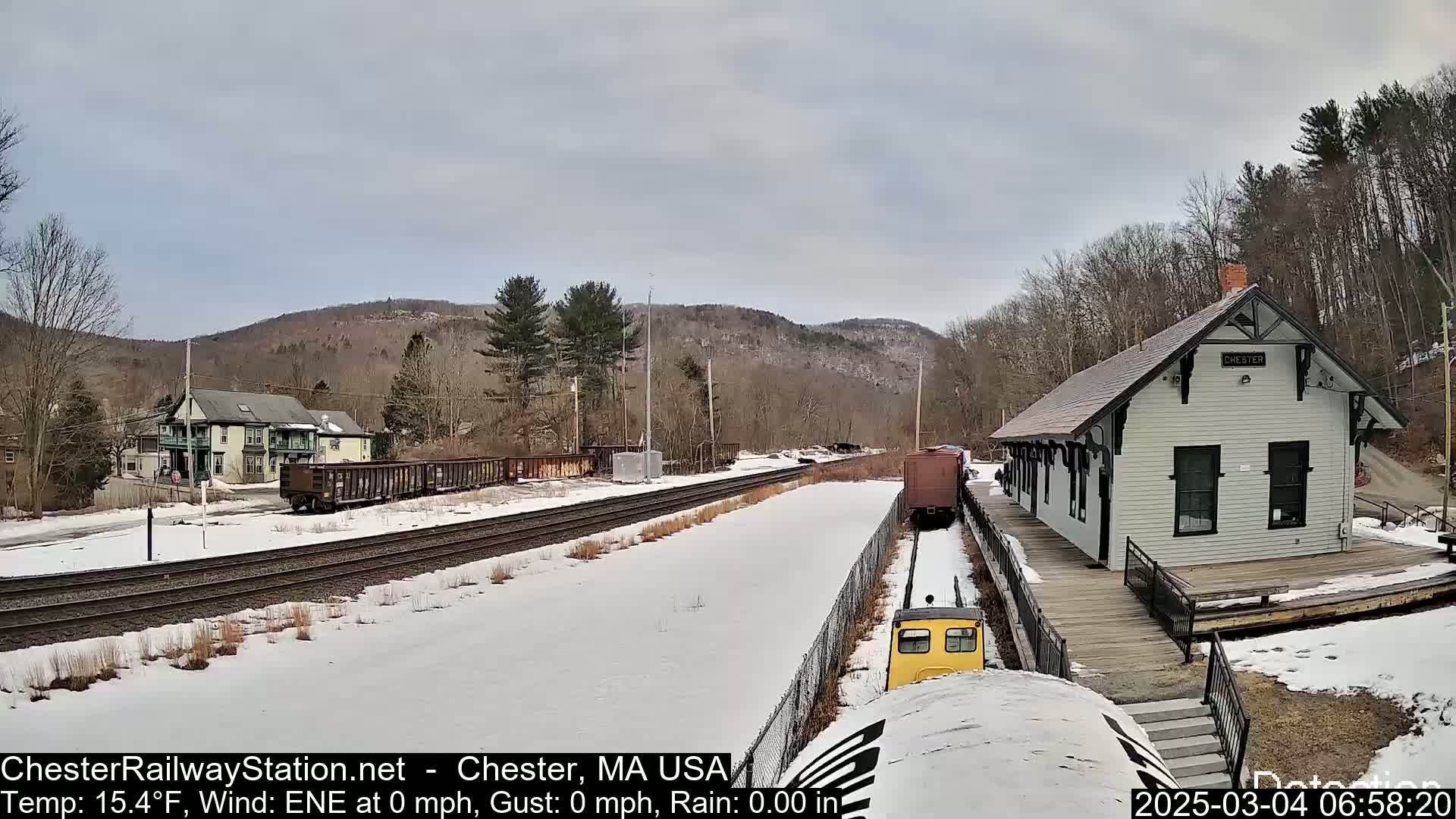 A small yellow train approaches a snow-covered train station next to a rail yard under a cloudy sky.