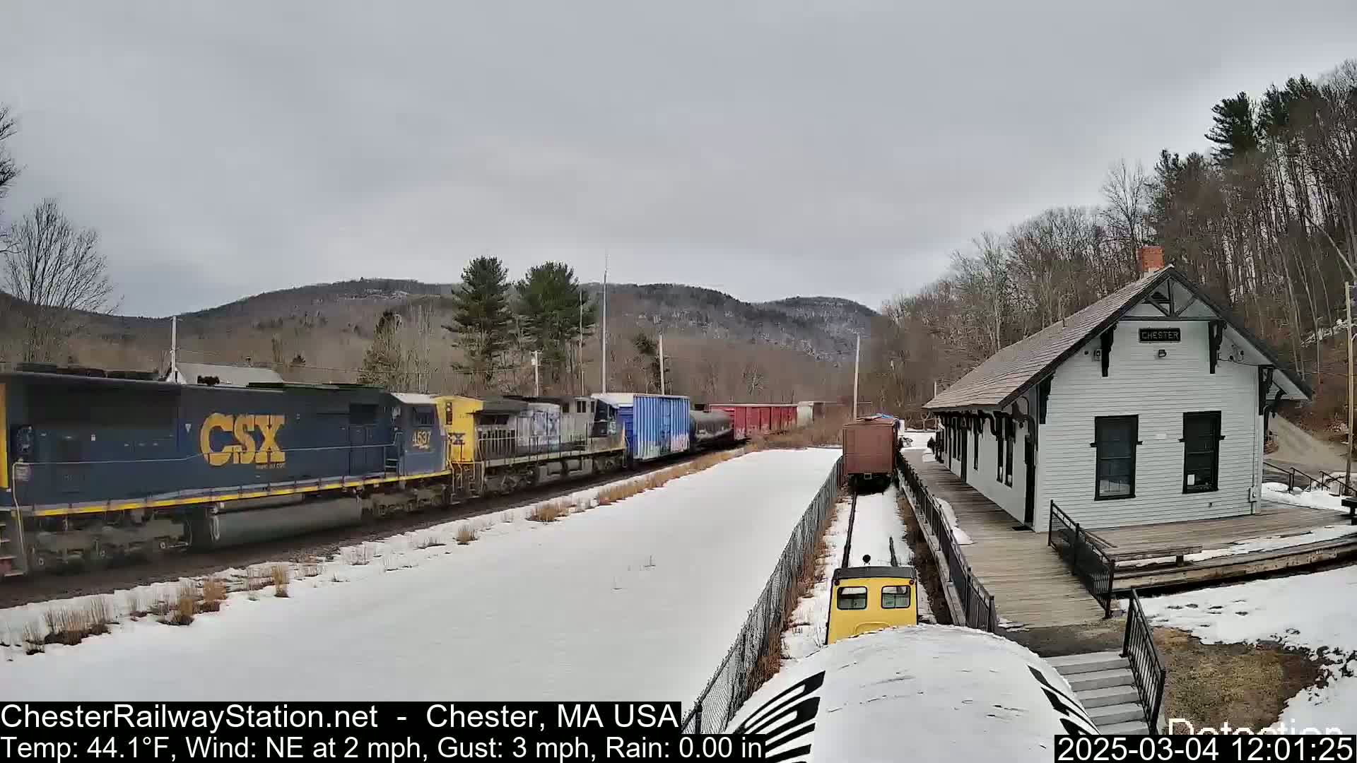 A long freight train passes a small, white train station on a snowy day with a northeast wind.