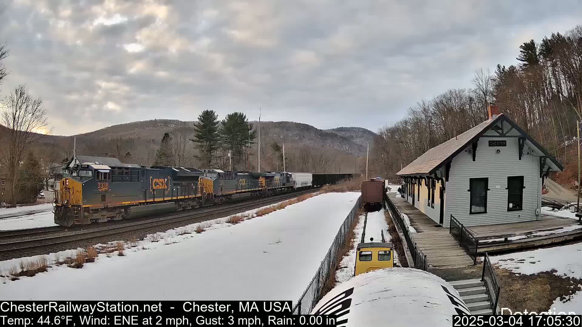 A freight train passes a snow-covered train station under a cloudy sky.