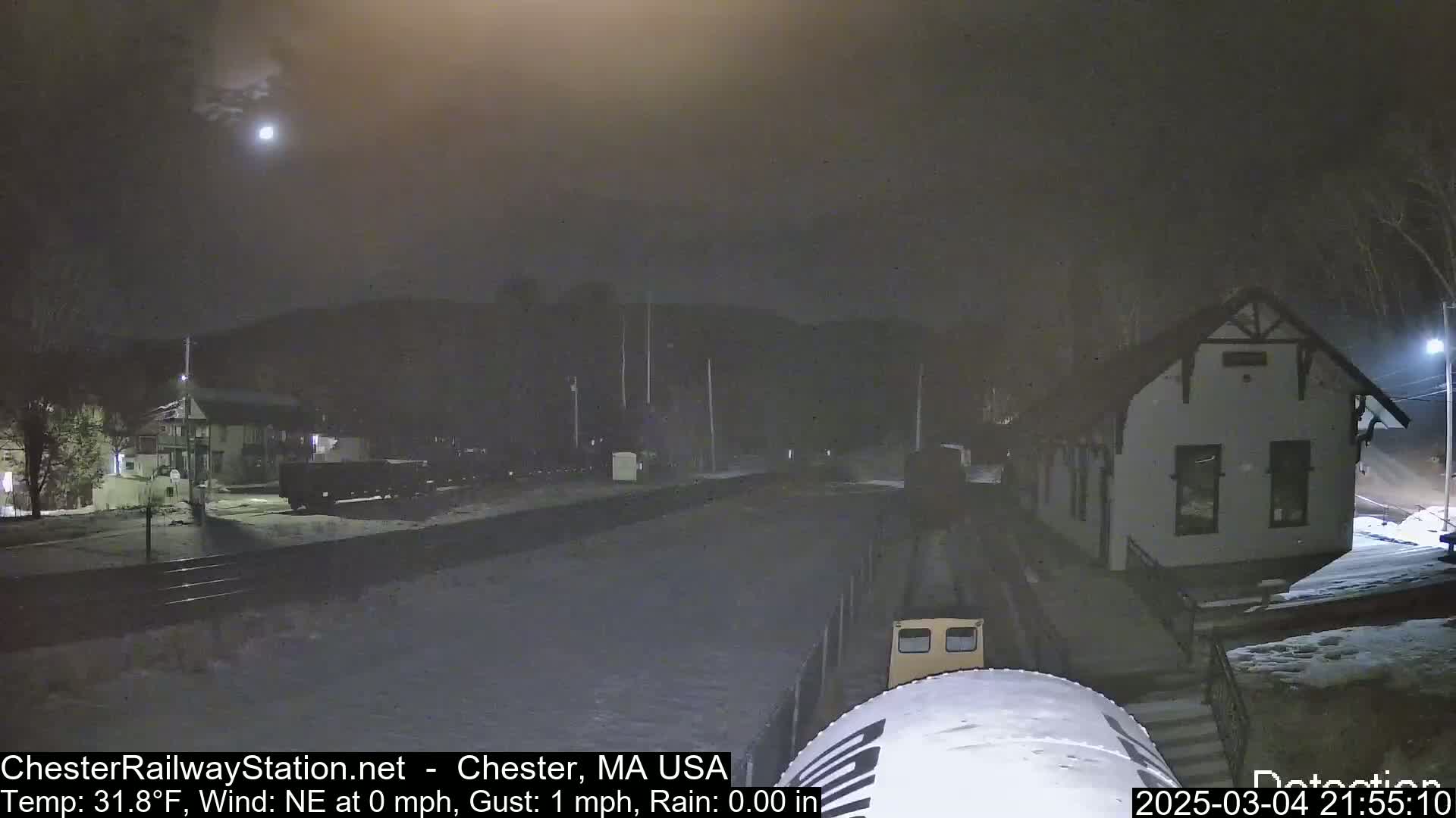 A nighttime view of a train at a snow-covered station under a dimly lit sky with a visible moon.