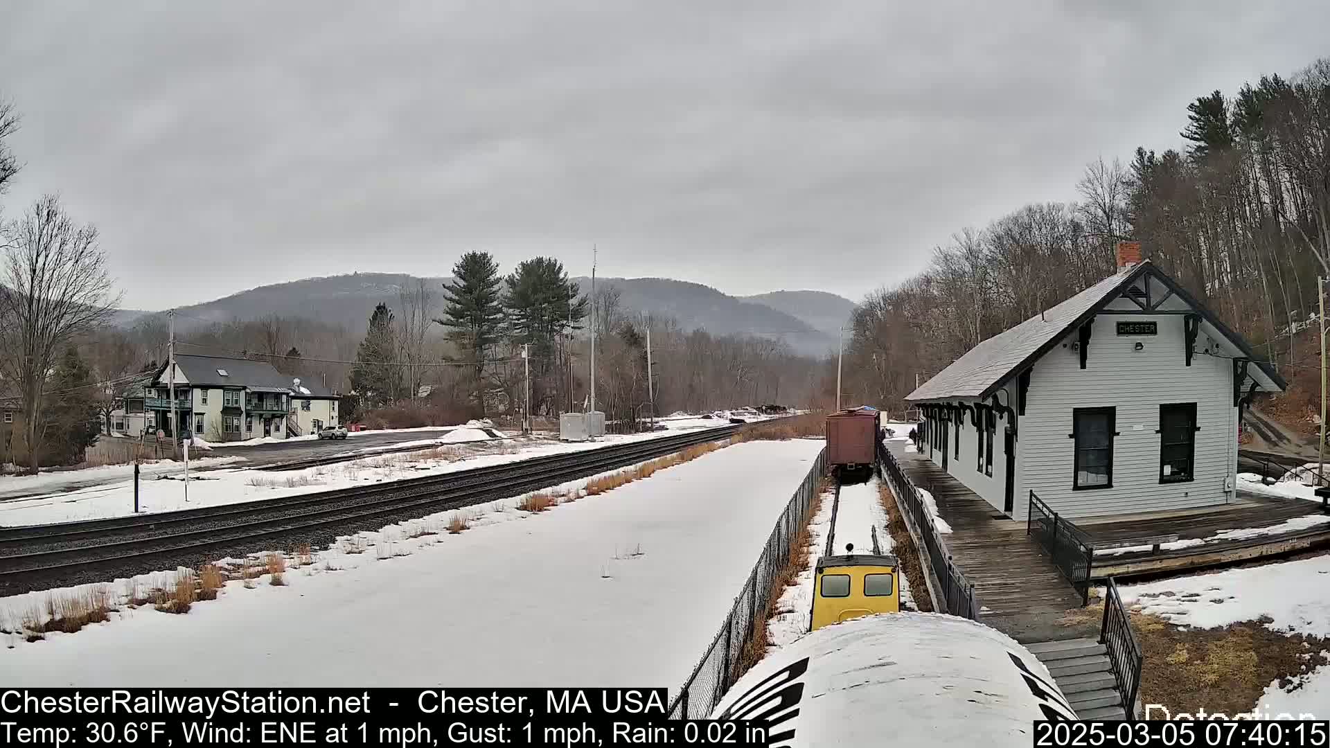 A snow-covered train station and railroad tracks are visible, with a small yellow train car and a freight car in the foreground, under a cloudy sky.