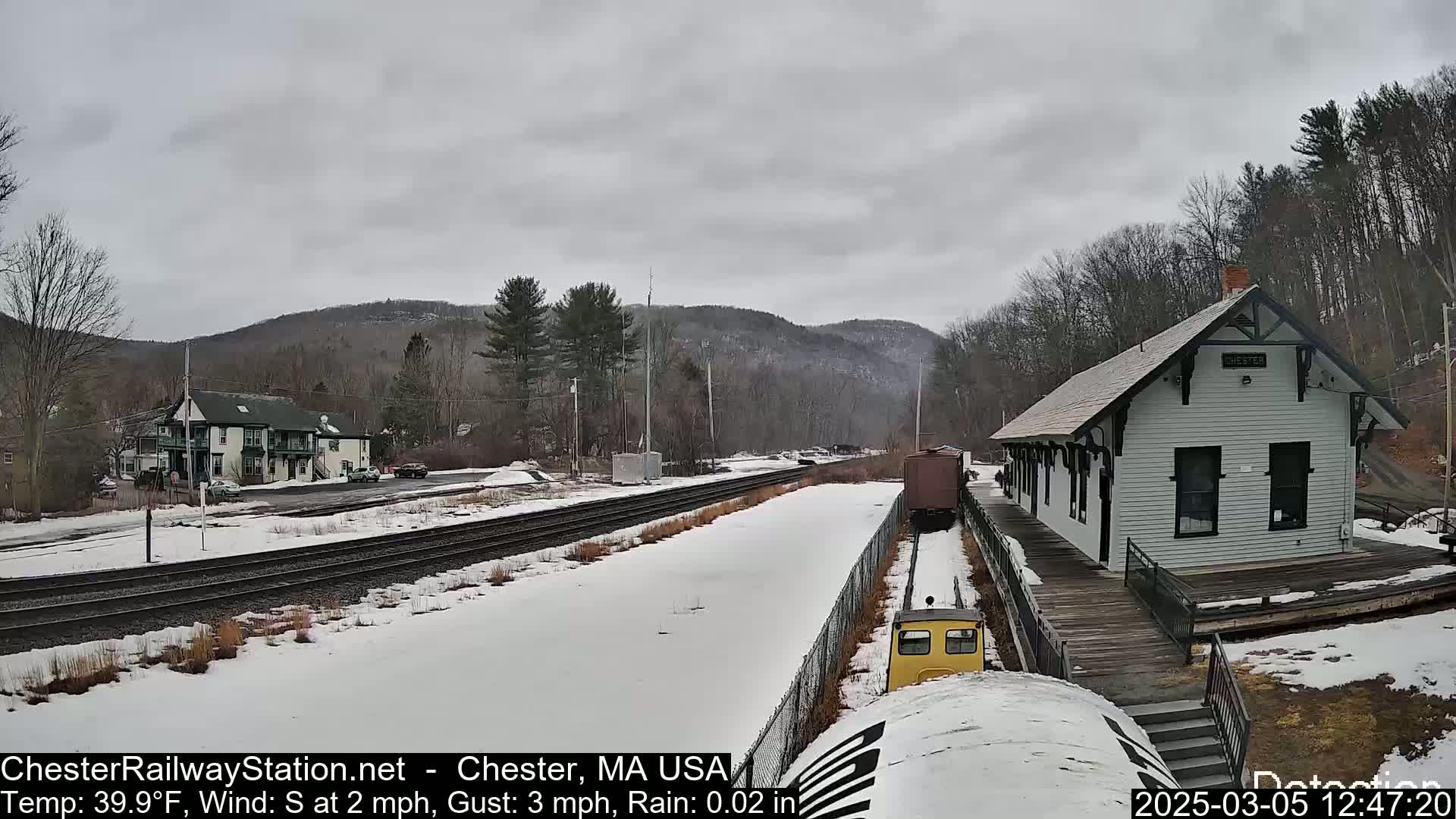 A snow-covered railway station and tracks are visible, with a small yellow train car and a larger building nearby, under an overcast sky.