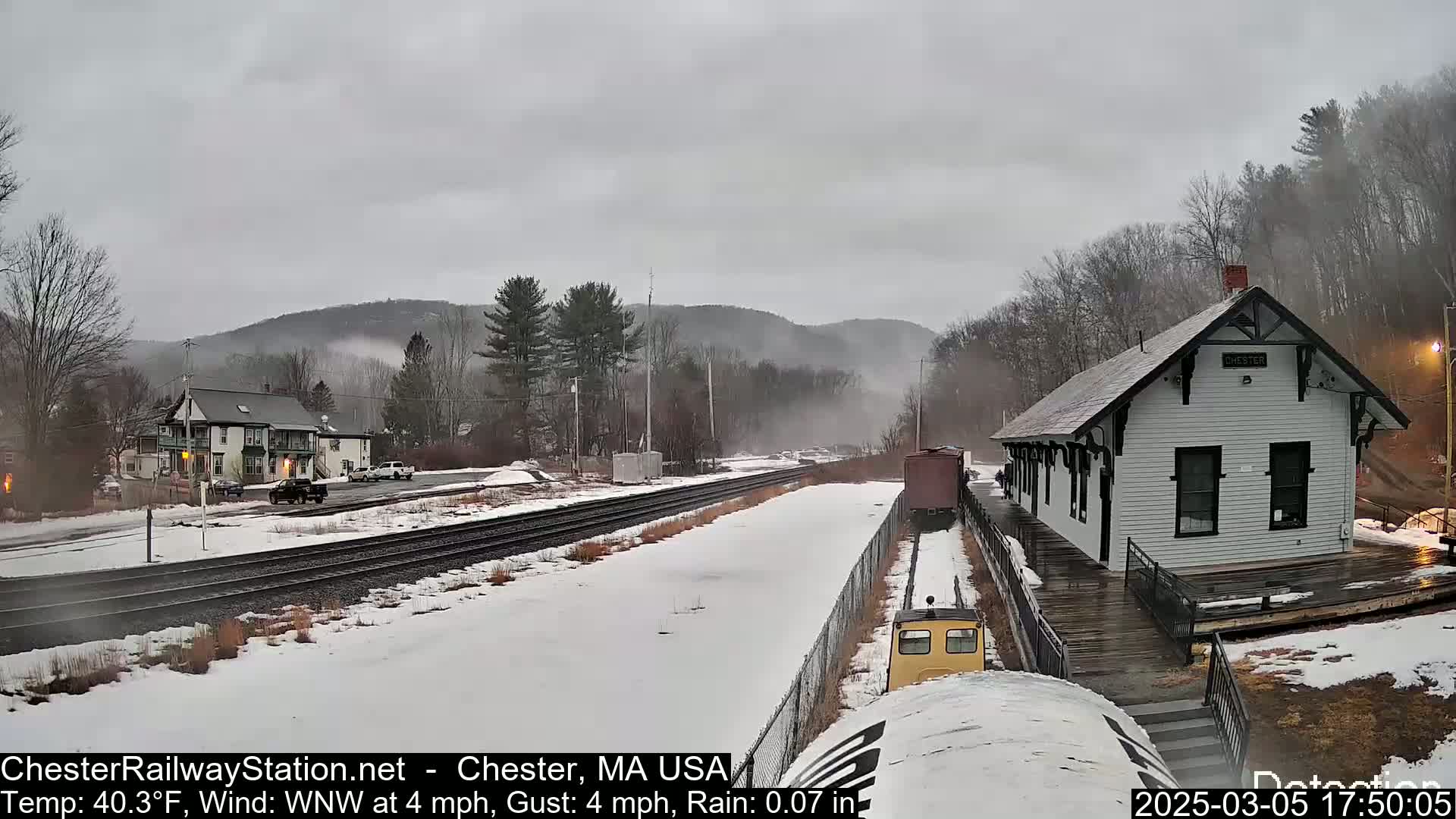 A snow-covered train station and railway tracks are visible under a gray, rainy sky, with misty hills in the background.
