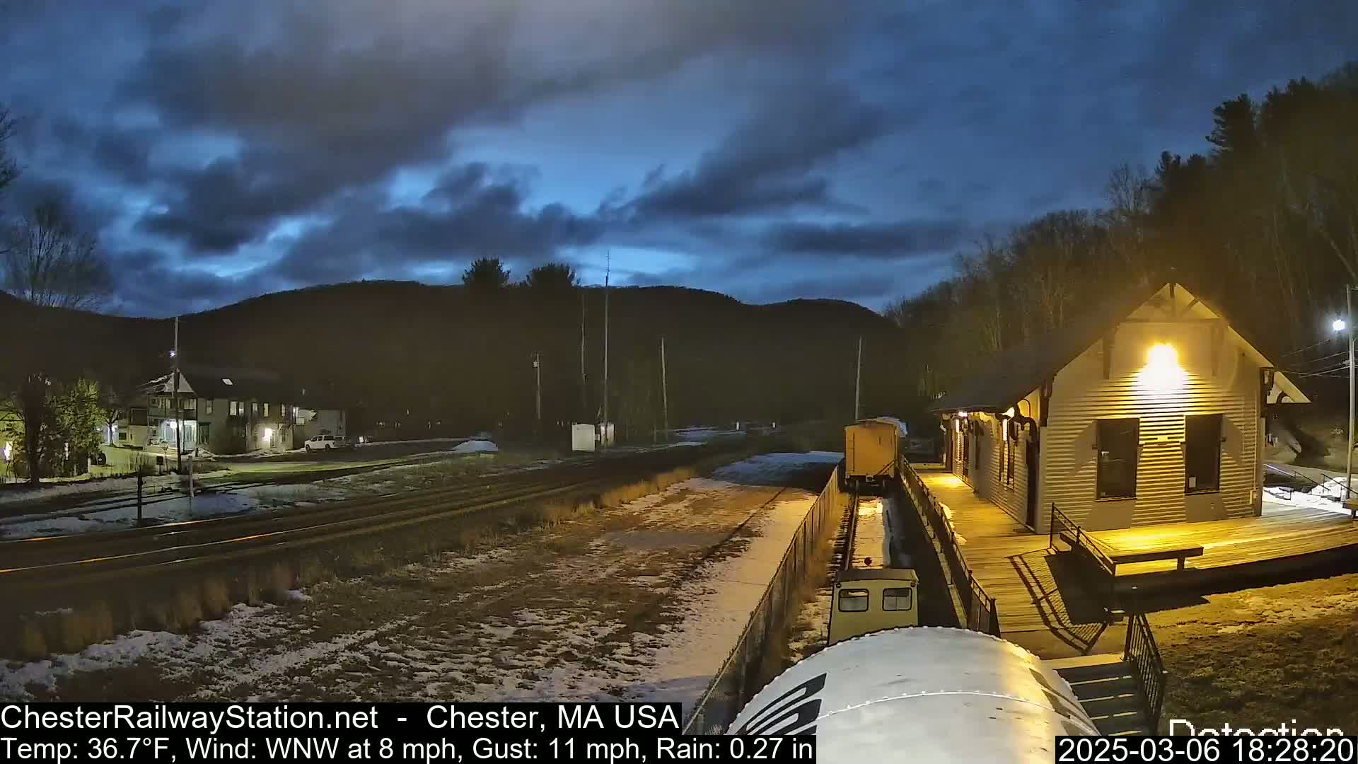 A train is approaching a lighted train station under a dark, cloudy sky with light rain and snow on the ground.