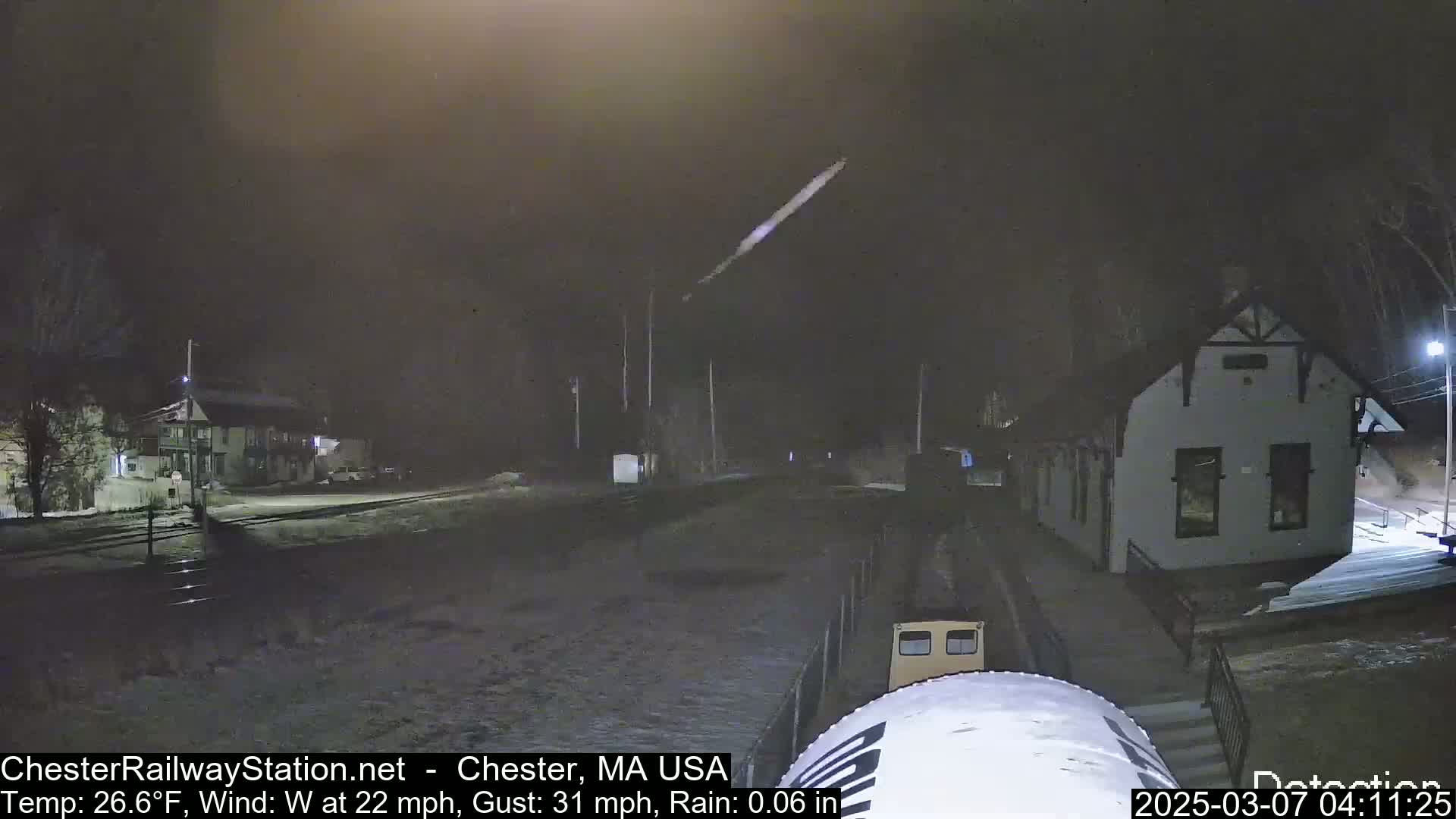 A train approaches a small building at night in rainy conditions with a 22 mph wind and gusts up to 31 mph.