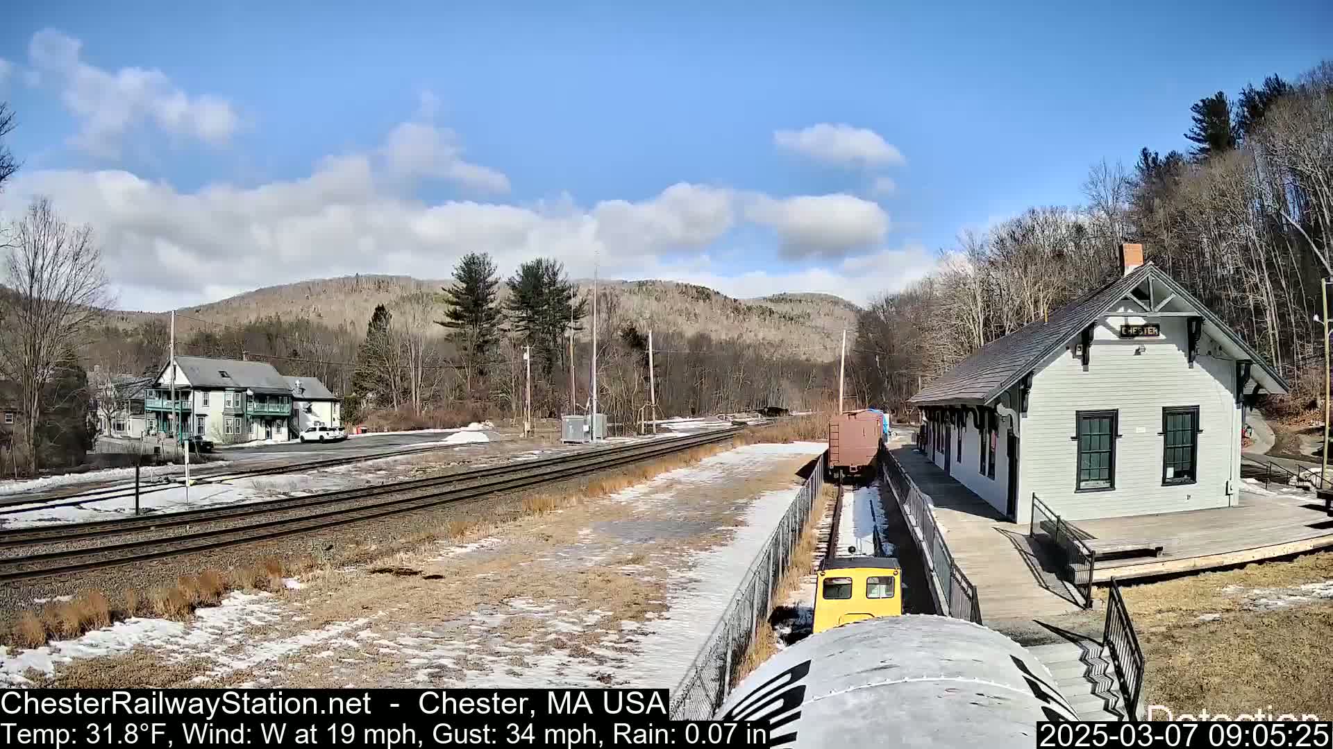 A small yellow train approaches a train station on a partly cloudy day with a light wind and some rain.