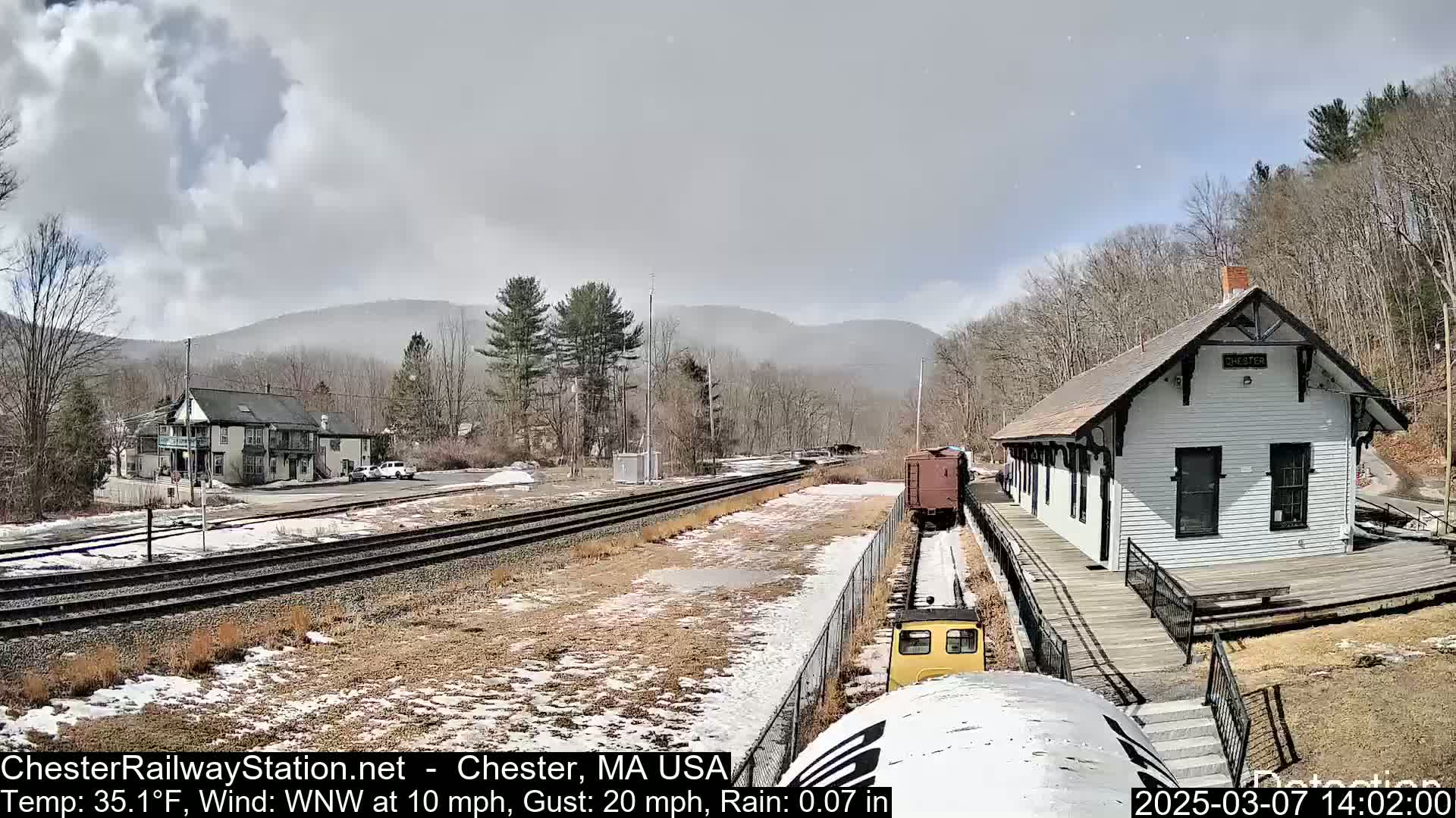 A train approaches a small, white train station in a snowy, partially cloudy landscape with a light rain and wind.