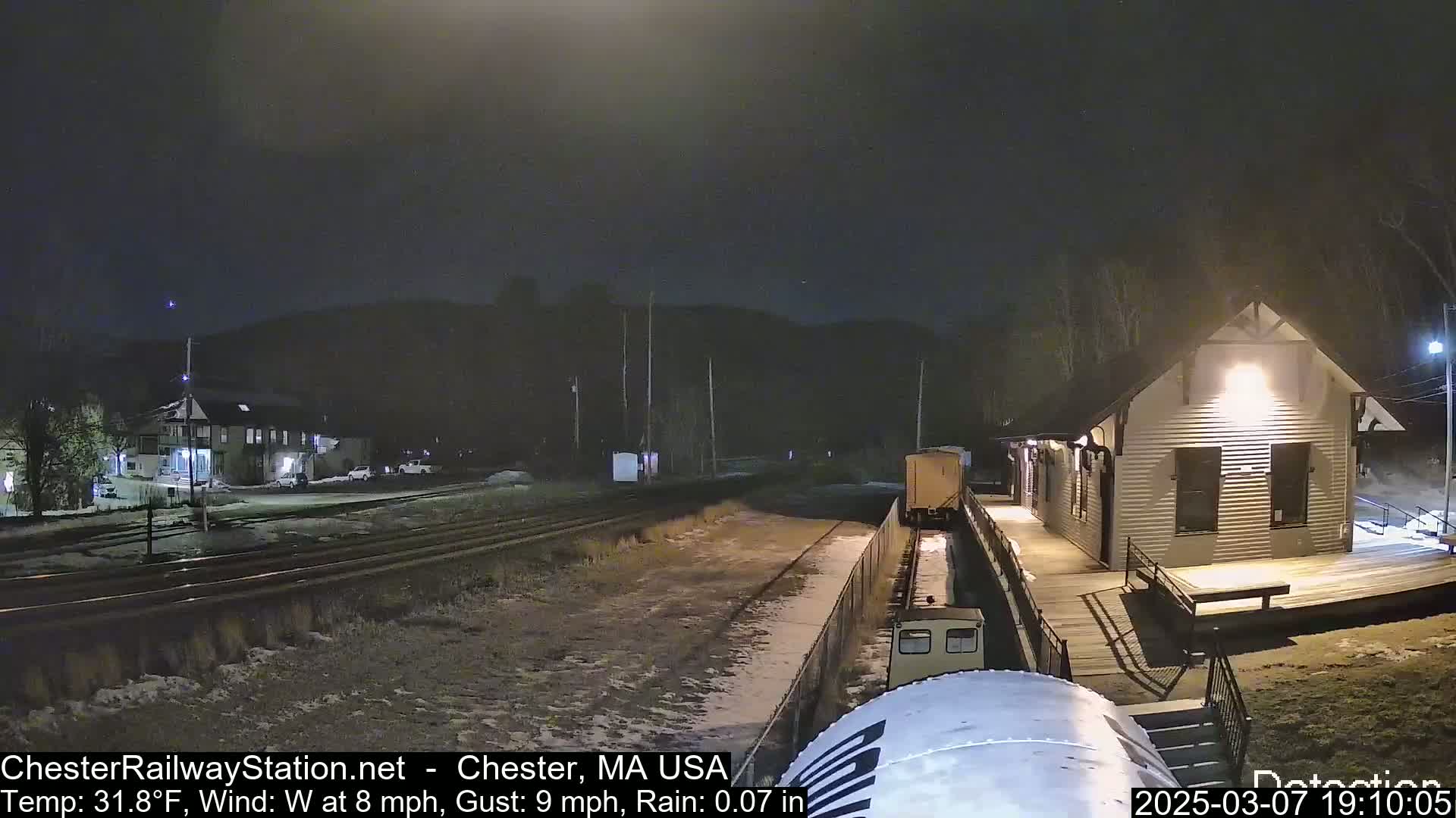 A nighttime view of a train arriving at a small, illuminated station, with light rain and snow on the ground.