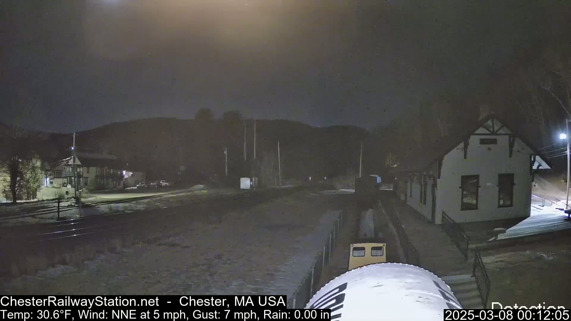 A nighttime view of a train approaching a small station building under a dark, cloudy sky.
