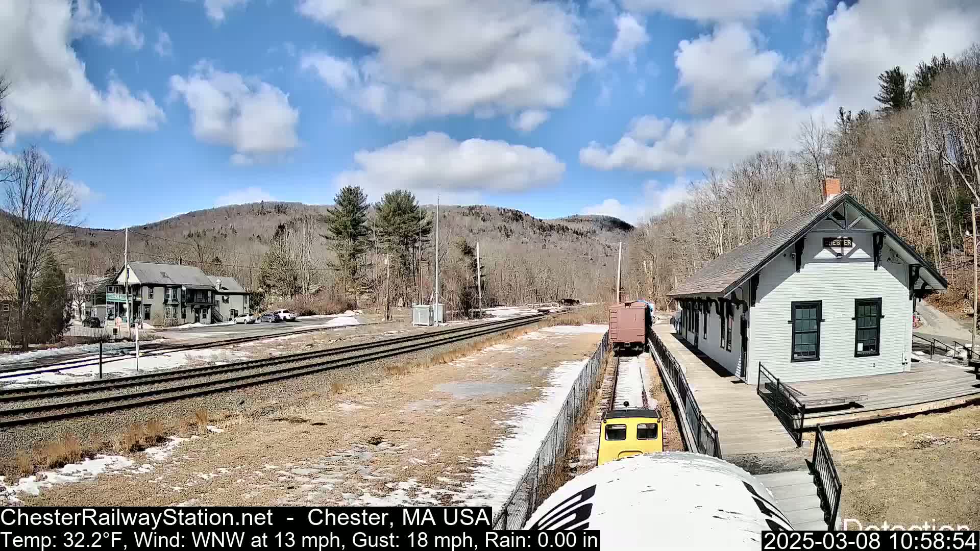 A small yellow train approaches a light gray train station on a partly sunny day with some patchy snow and ice on the ground,  set against a backdrop of leafless trees and hills.