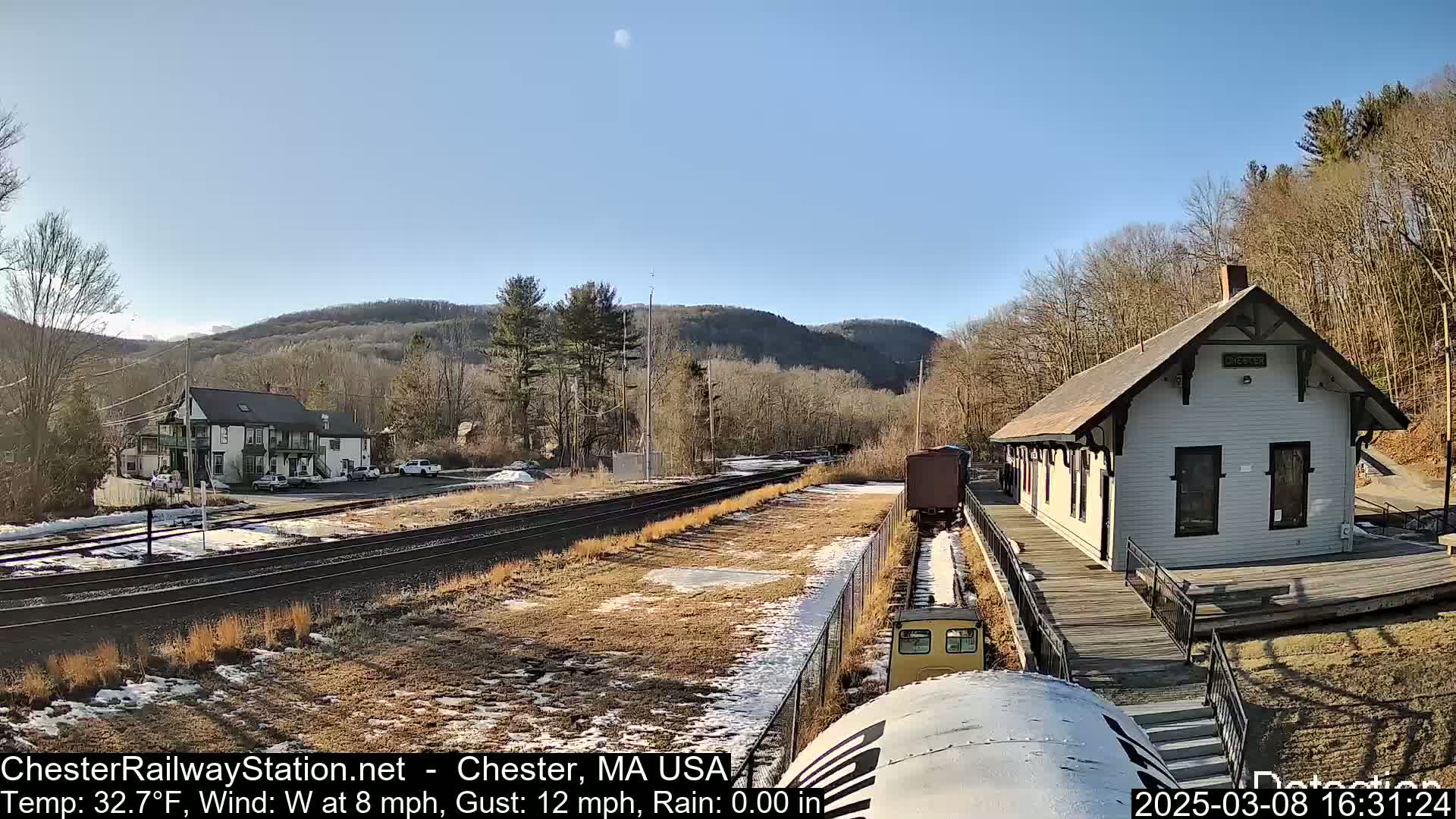 A train approaches a small, white train station situated beside railroad tracks, with a building and wooded hills visible in the background, under a clear, sunny sky.