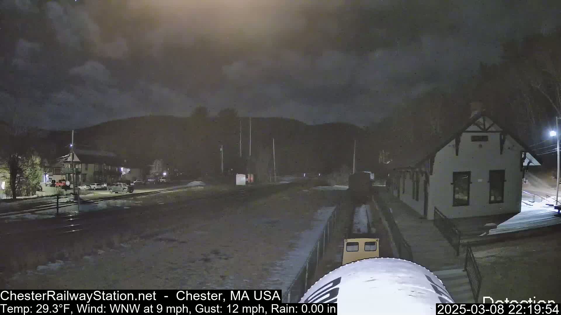 A nighttime view of a train station and railroad tracks under a cloudy sky, with some snow or ice on the ground.