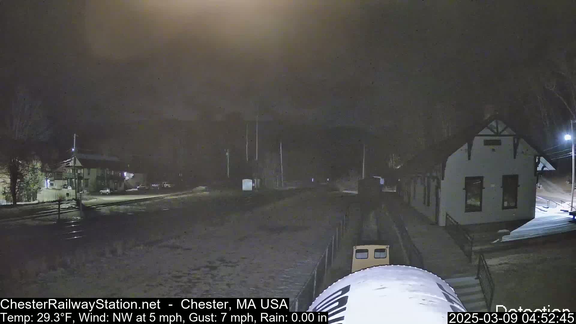 A nighttime view of a train approaching a station in snowy conditions, with a building and some parked cars visible in the background.