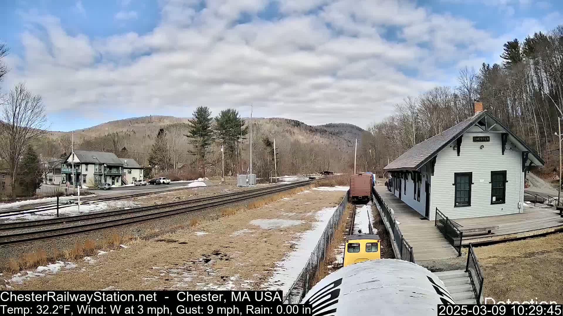 A small, white train station with a wooden platform sits beside train tracks on a partly cloudy day, with a small train approaching and a view of bare trees and hills in the background.