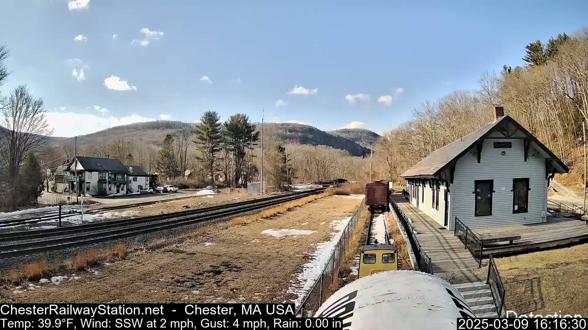 A train approaches a small, white train station nestled amongst bare trees and hills under a mostly sunny sky.