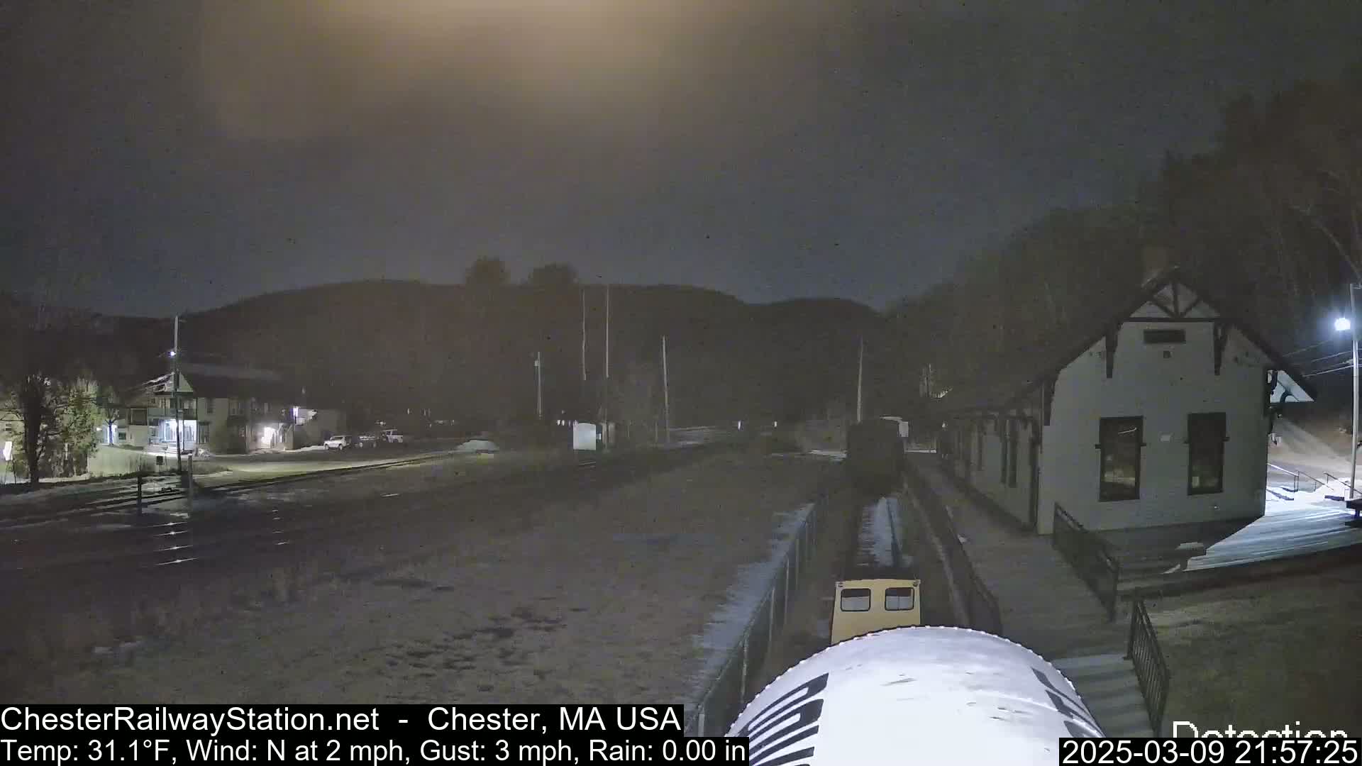A nighttime view of a train approaching a station, with light snow on the ground and a dark, overcast sky.